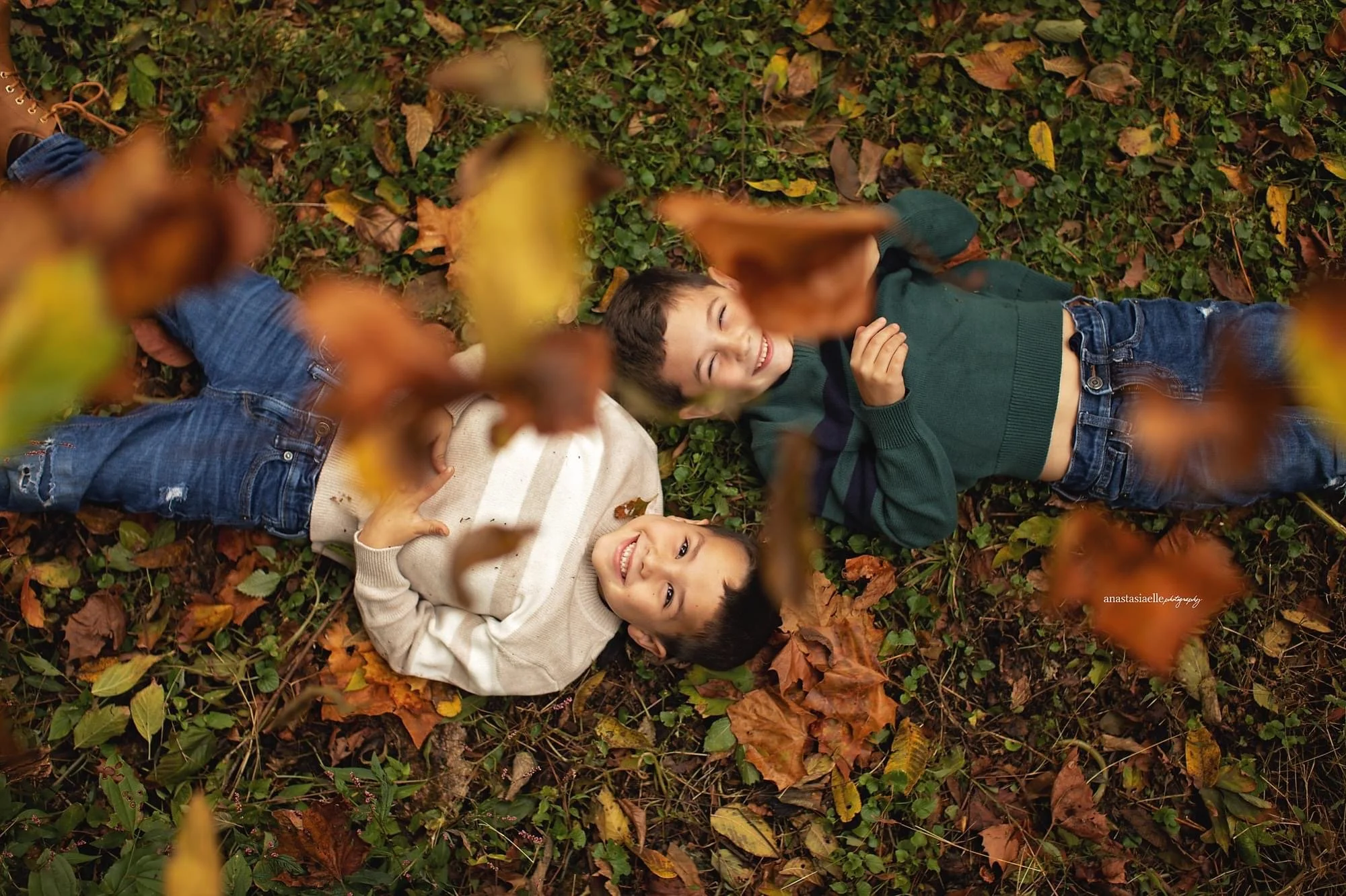 Two young boys lying on the ground covered with fallen autumn leaves, smiling and looking up at the camera.