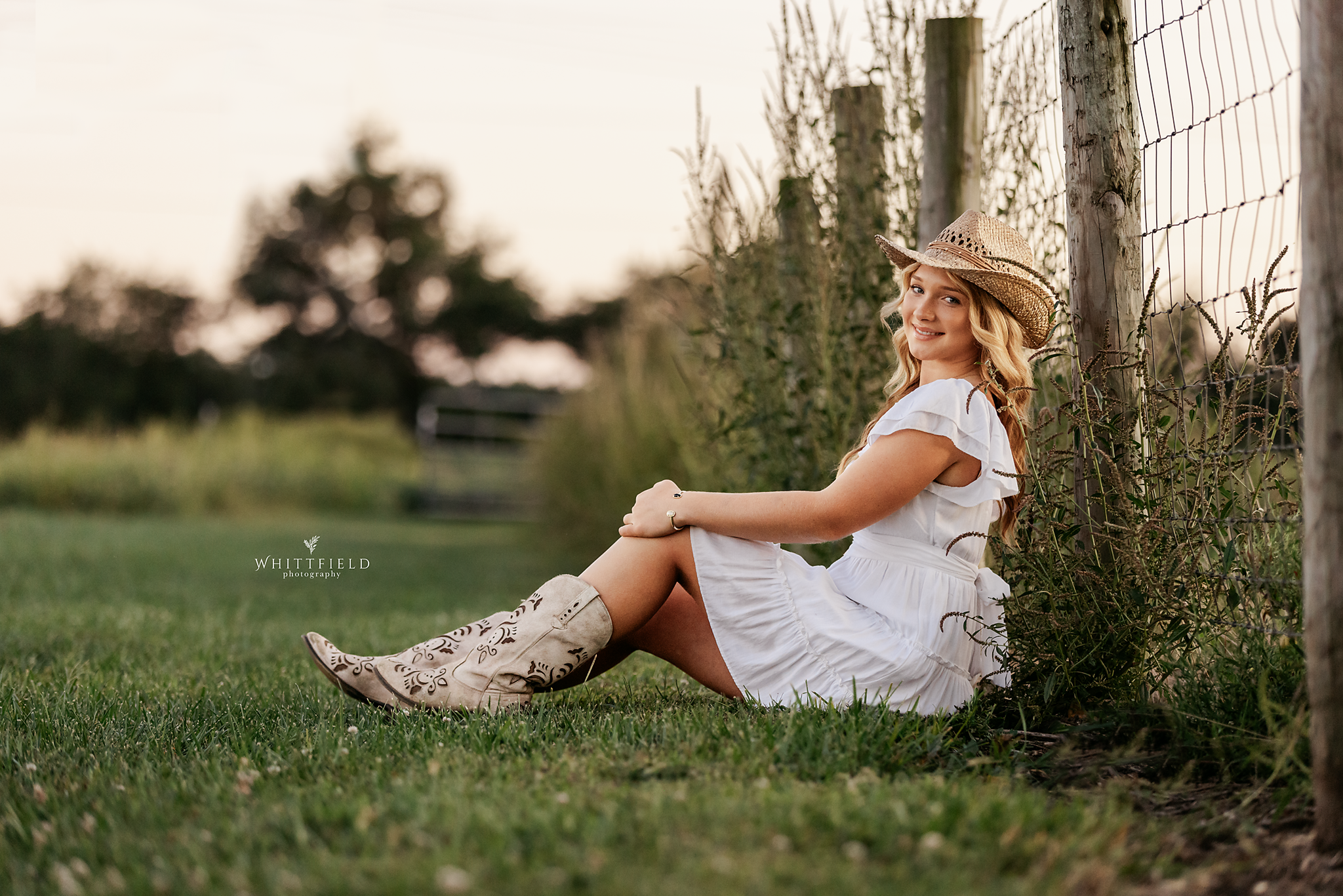 A young woman with blonde hair, wearing a white dress and cowboy boots, sitting on grass beside a wooden fence, smiling at the camera during sunset.
