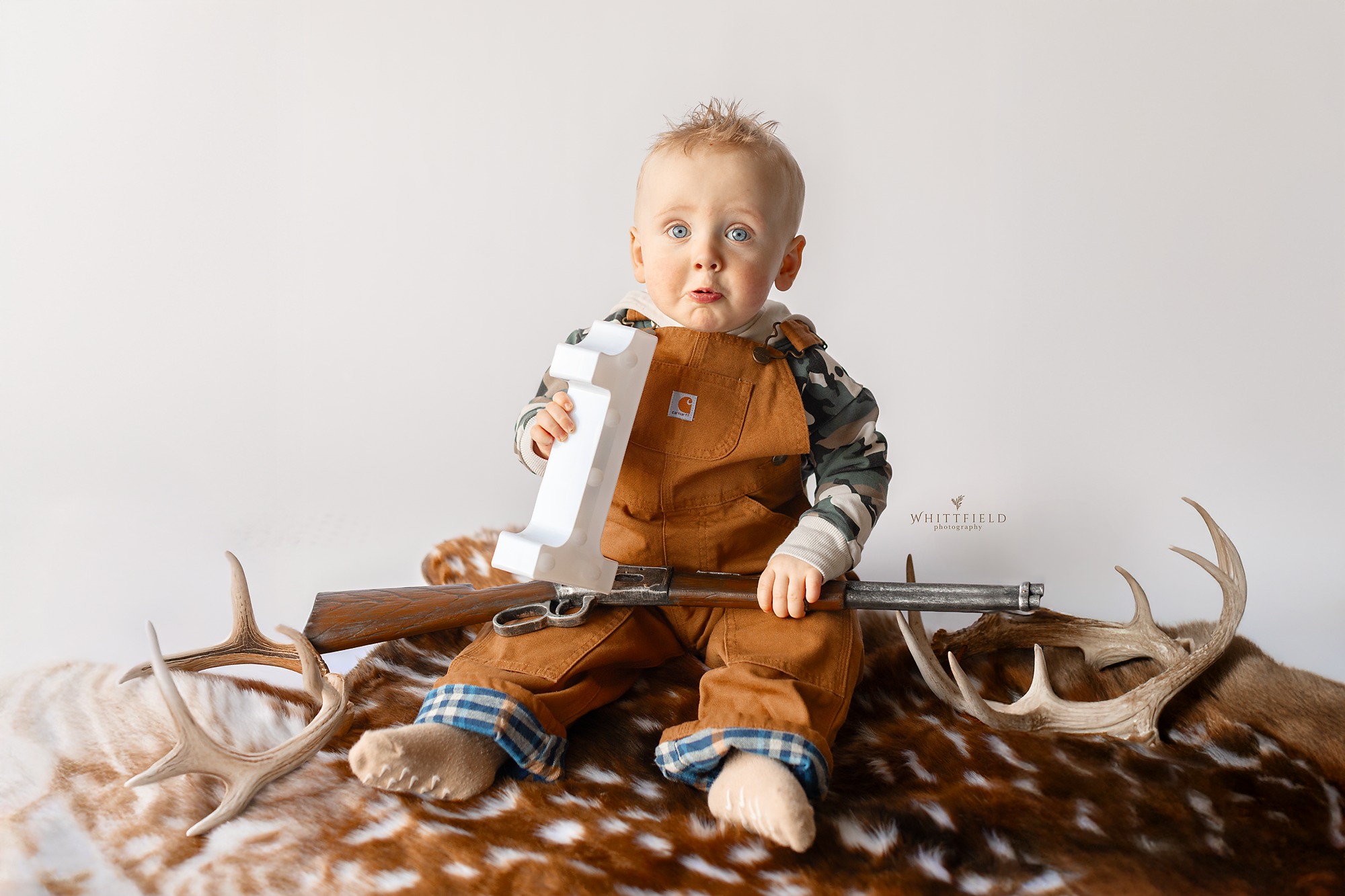 A young boy with blue eyes and blond hair sits on a brown and white animal fur, holding a toy rifle and a white plastic number one. Decorative antlers are placed around him, and he is dressed in a camouflage shirt, brown overalls, and beige socks. Th