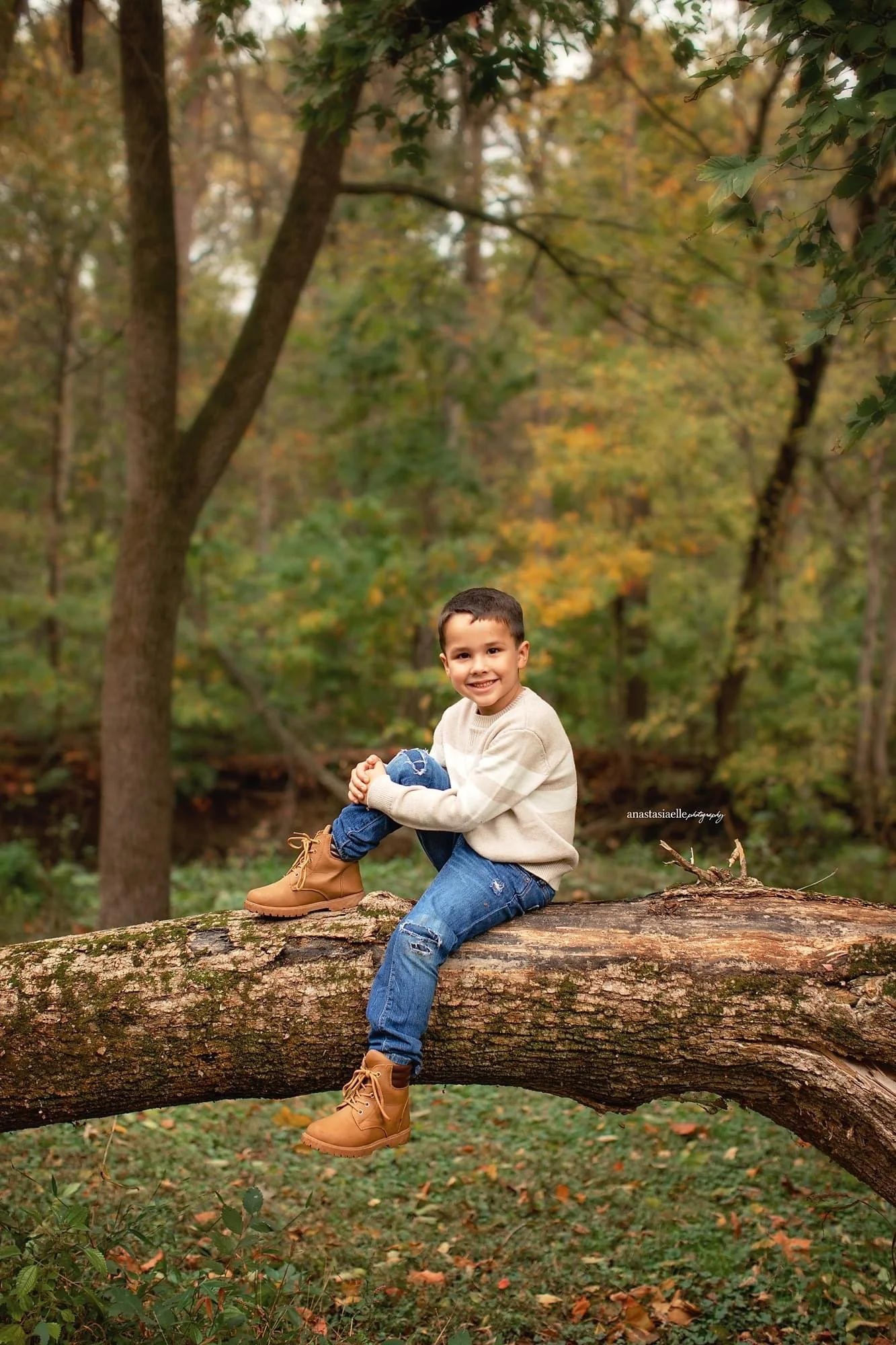A young boy smiles while sitting on a fallen tree trunk in a forest with autumn foliage.