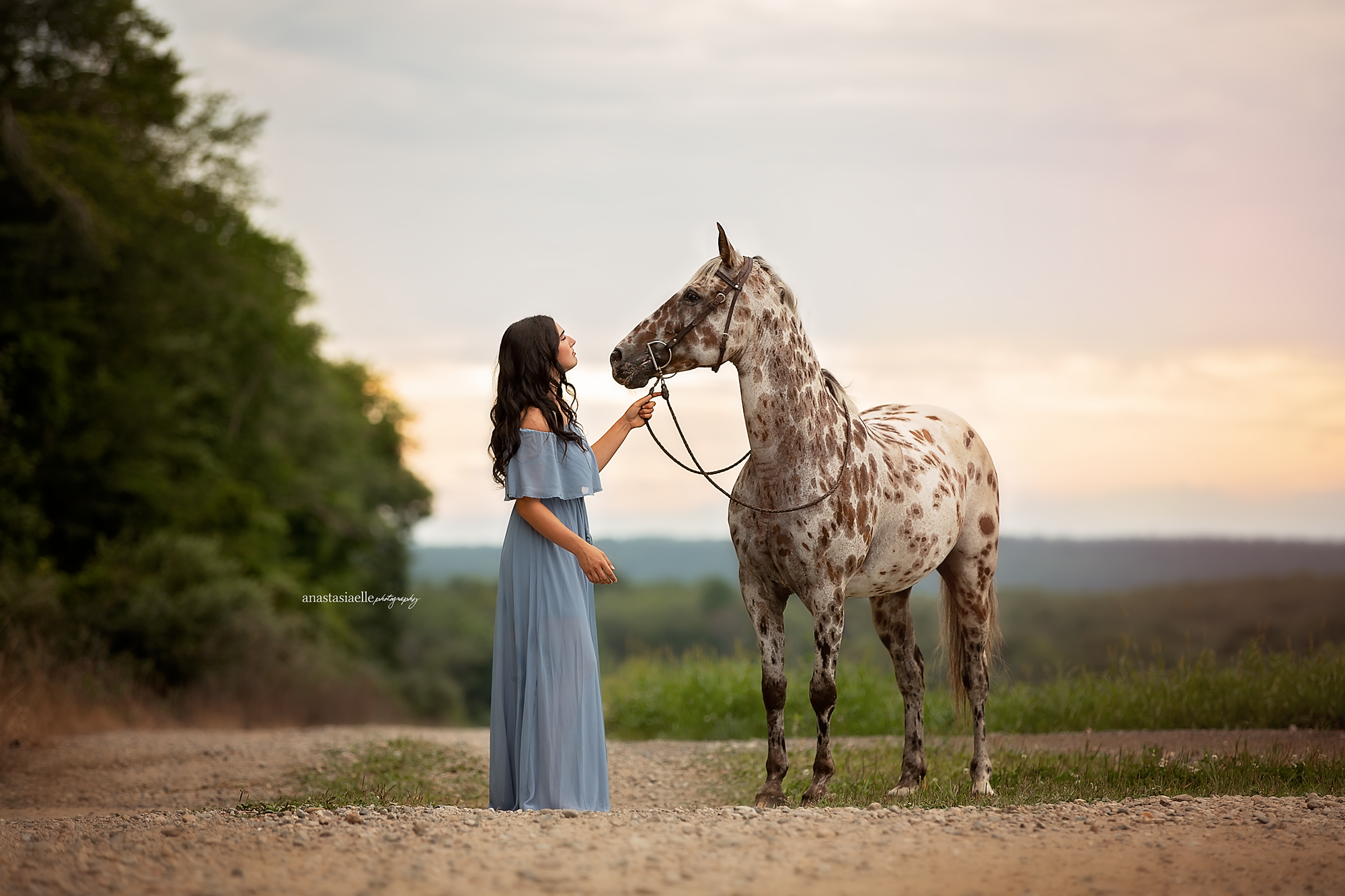 A woman with long dark hair in a blue dress holding a leash of a gray and white spotted horse on a dirt path during sunset.