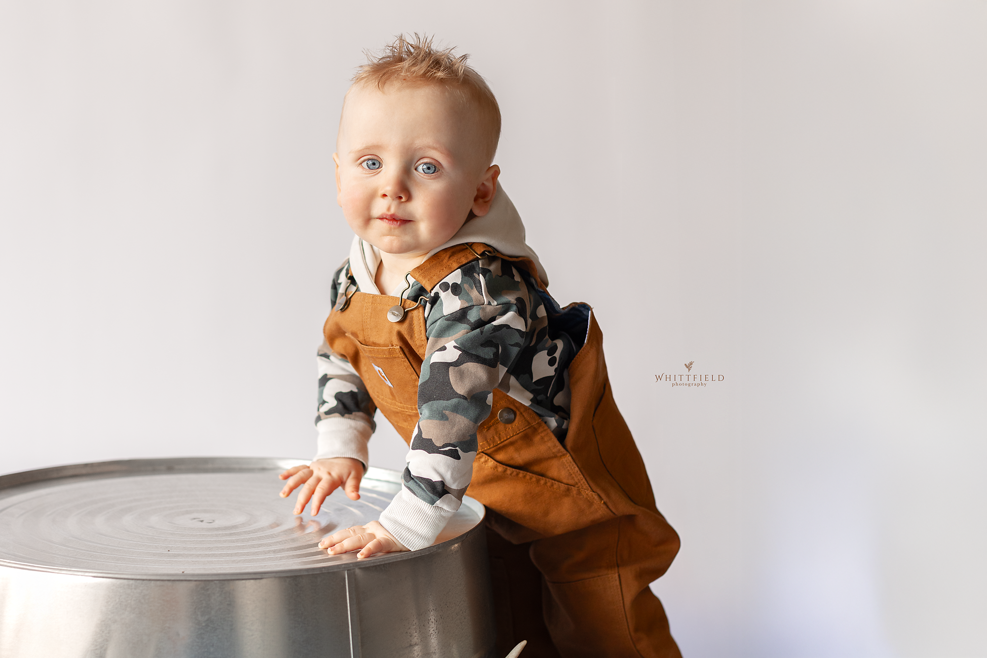 Cute toddler with blue eyes, blonde hair, wearing camouflage hoodie and brown overalls, standing and leaning on a round metallic table against a plain white background.