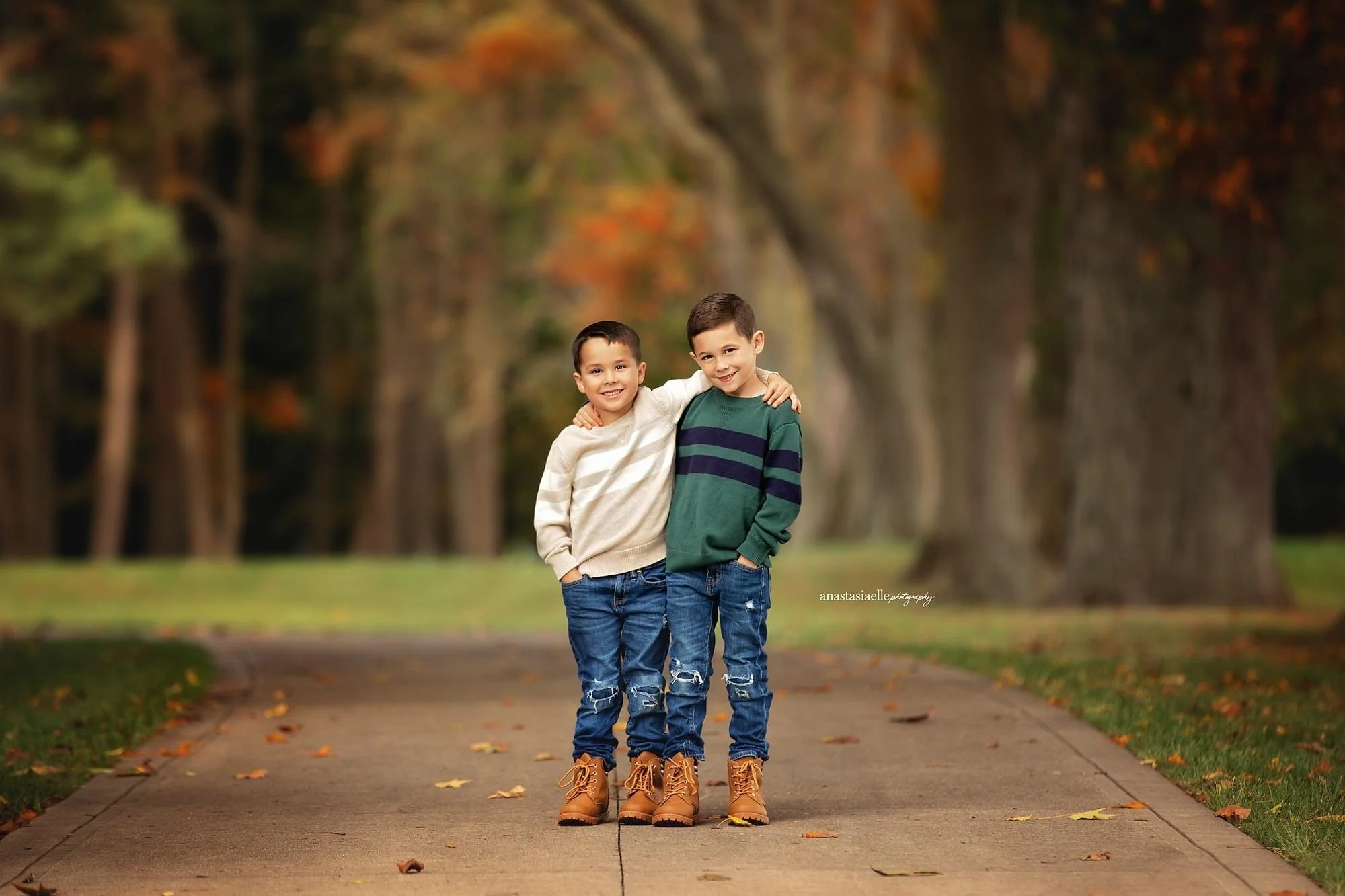 Two young boys standing on a park sidewalk surrounded by fall trees, dressed warmly in sweaters, jeans, and boots, smiling and hugging each other.