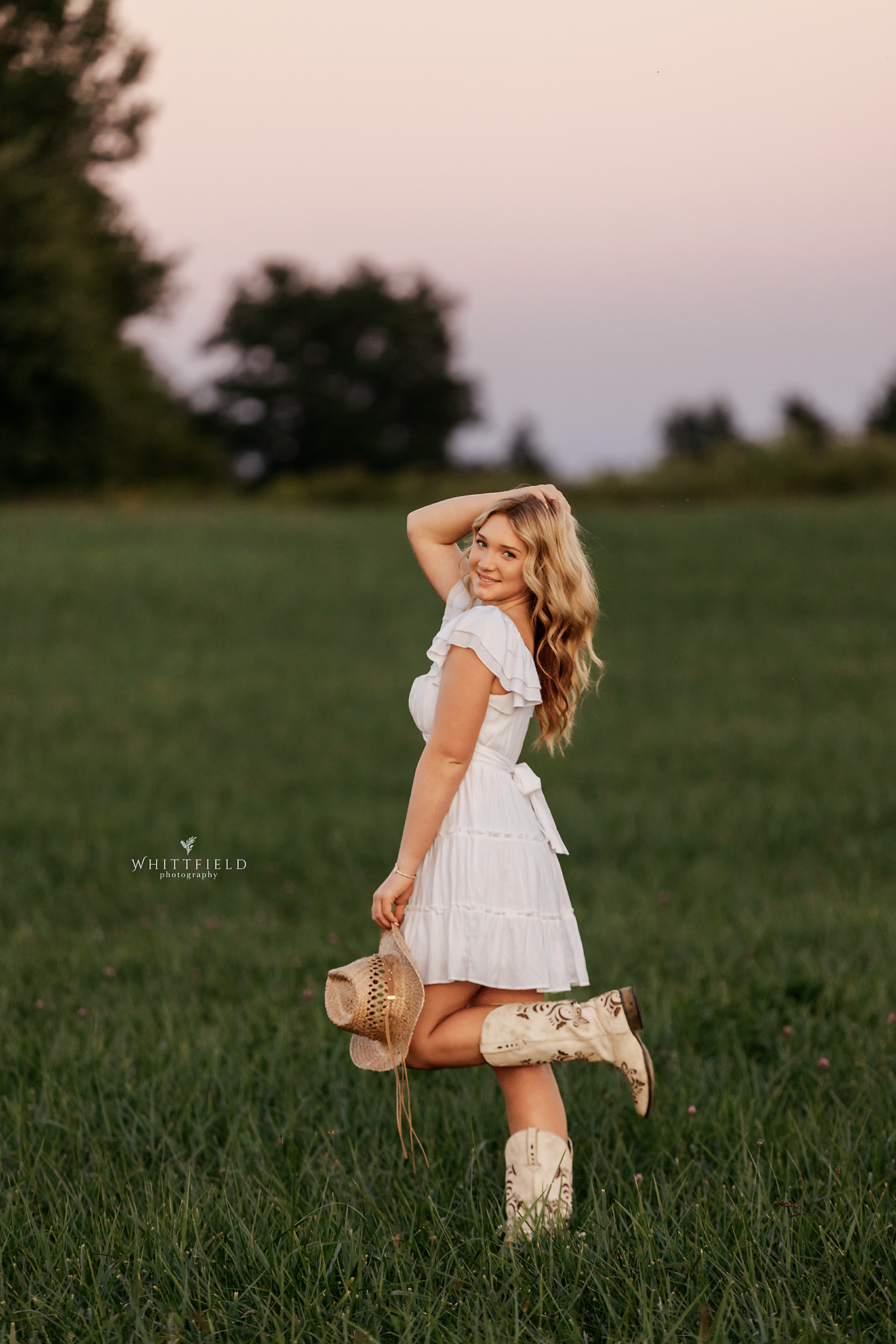 A young woman in a white dress and cowboy boots standing in a grassy field at sunset, holding a straw hat, smiling and posing with one hand behind her head.