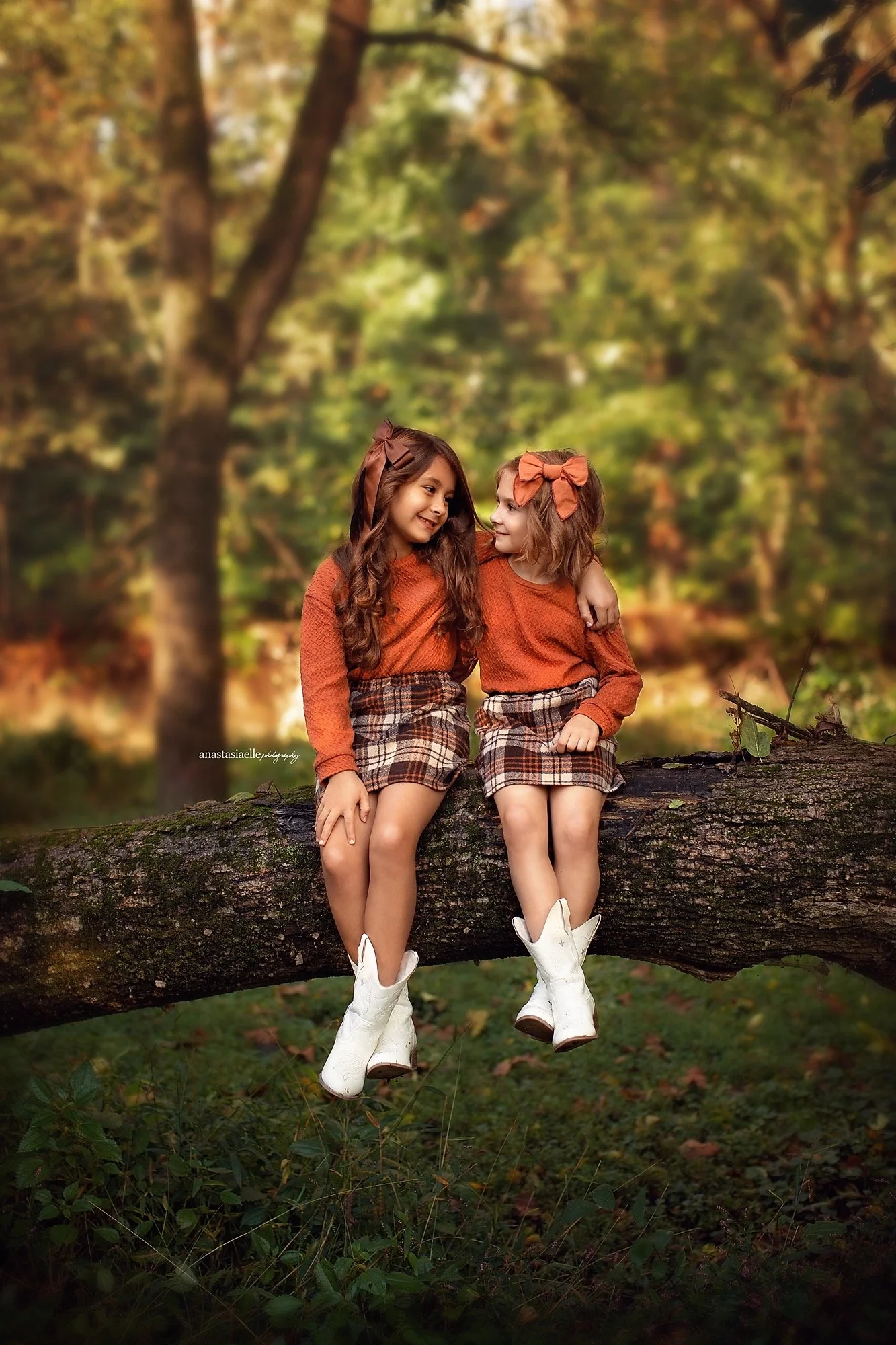 Two young girls with brown and blonde hair, wearing matching plaid skirts, orange sweaters, and white cowboy boots, sitting on a fallen tree trunk in a forest during autumn, smiling at each other.