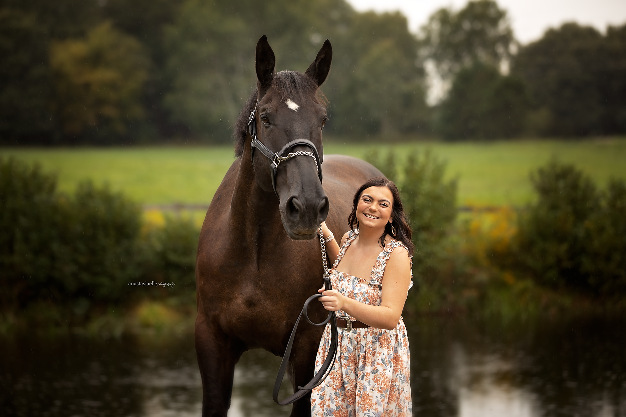 A woman standing next to a horse outdoors in a green field and water, smiling at the camera.