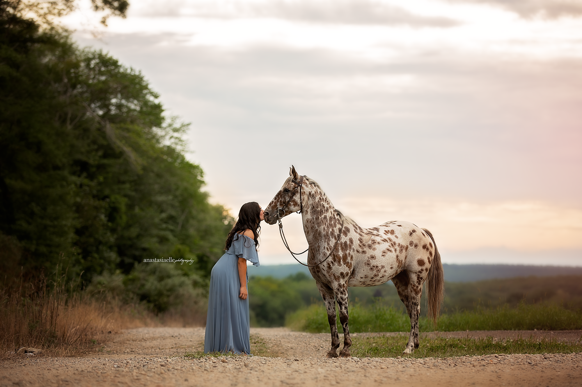 A woman in a long, flowing blue dress kisses a spotted horse on a dirt path amidst trees and open landscape during sunset.