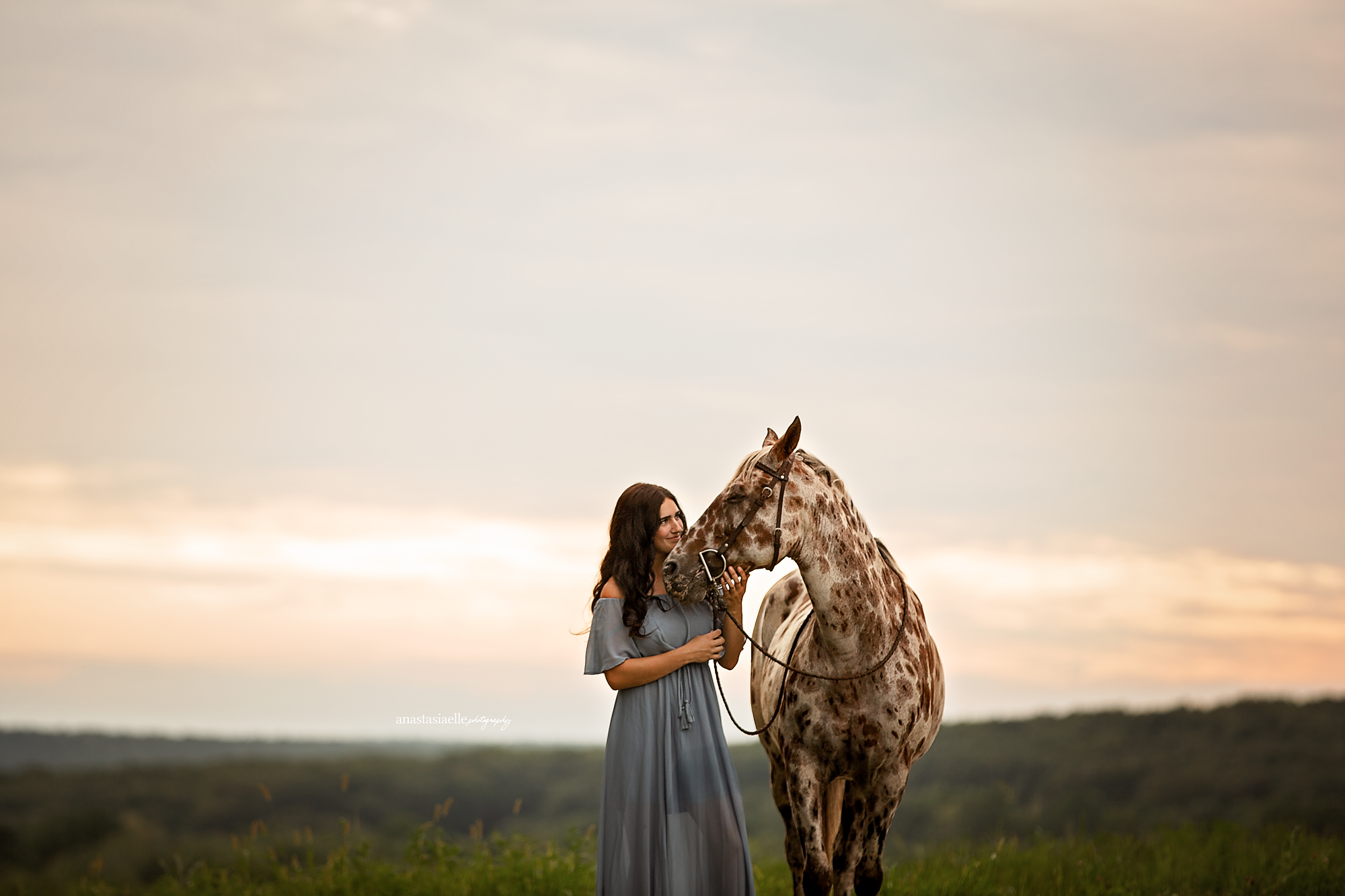 A woman in a long, off-the-shoulder gray dress is standing next to a tall, spotted giraffe in an open field at sunset.