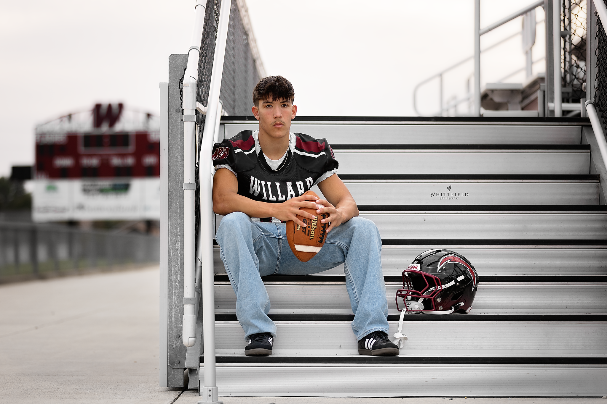 A young male football player in a black Willard jersey sitting on stadium bleachers holding a football, with a black and red football helmet placed beside him on the steps.