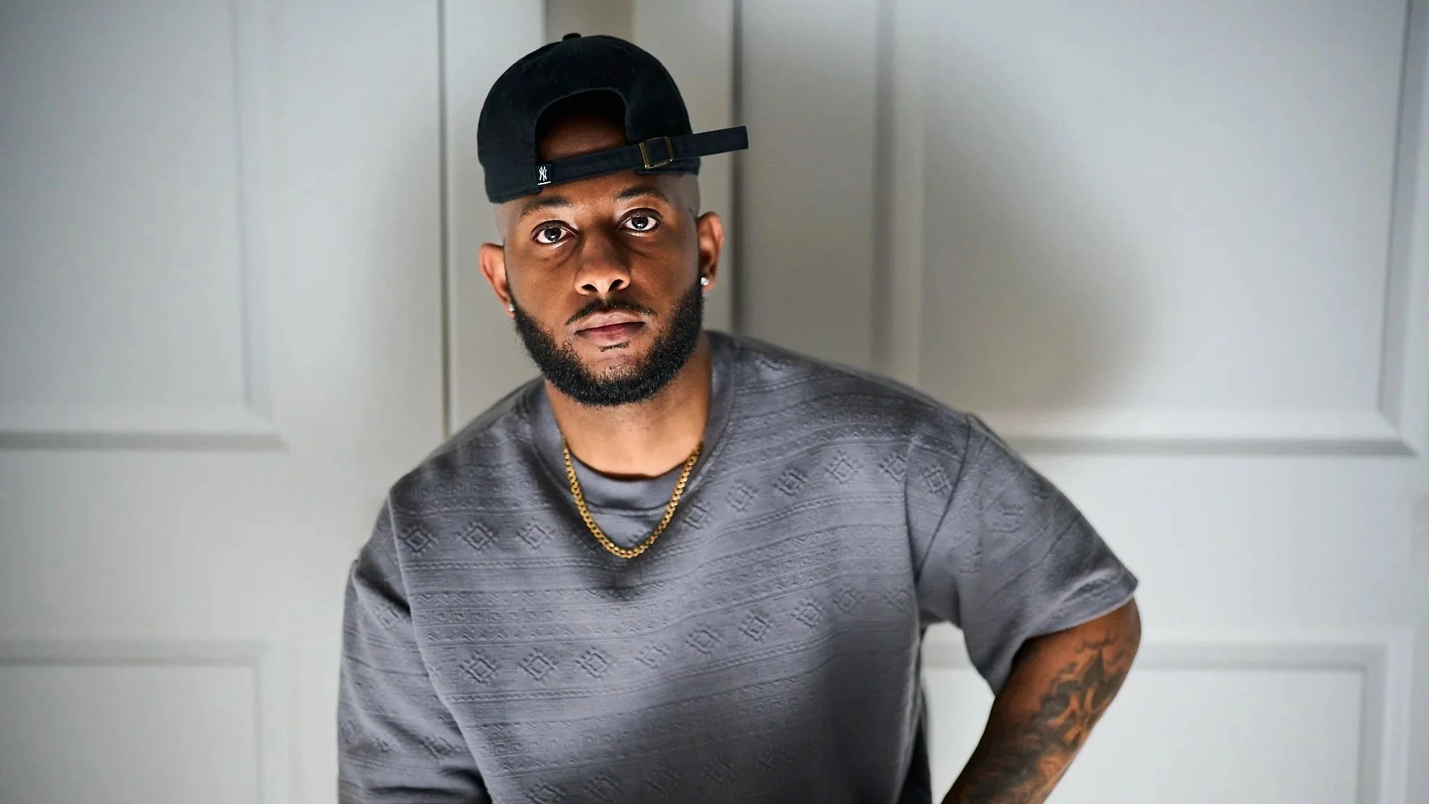 A young Black man wearing a black cap backwards, a gray patterned t-shirt, a gold chain, and earrings, standing against a neutral background with a serious expression.