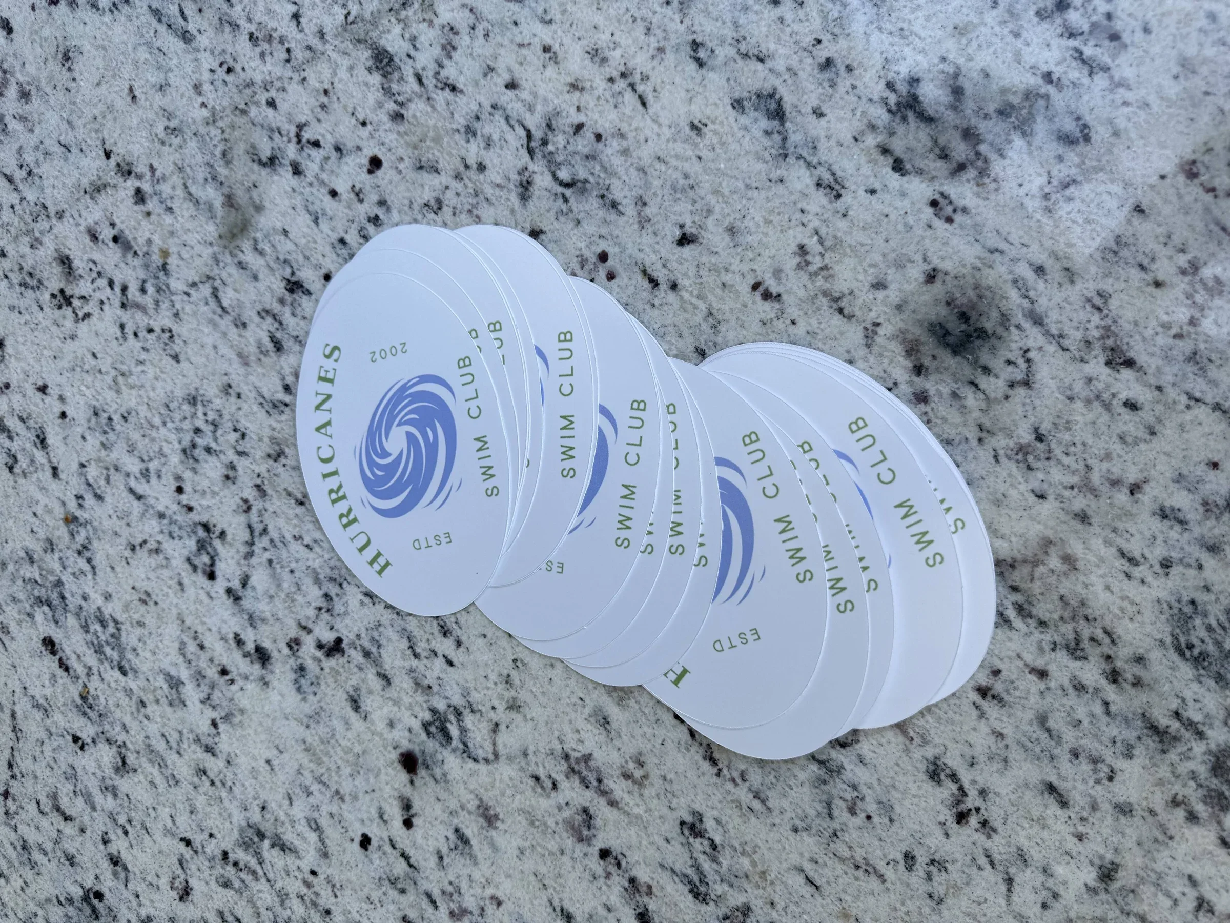 Stacked round holographic stickers with blue swirl logo and green text, reading 'HURRICANES', 'SWIM CLUB', and 'ESTD 2002', on a rough, light-colored stone surface.