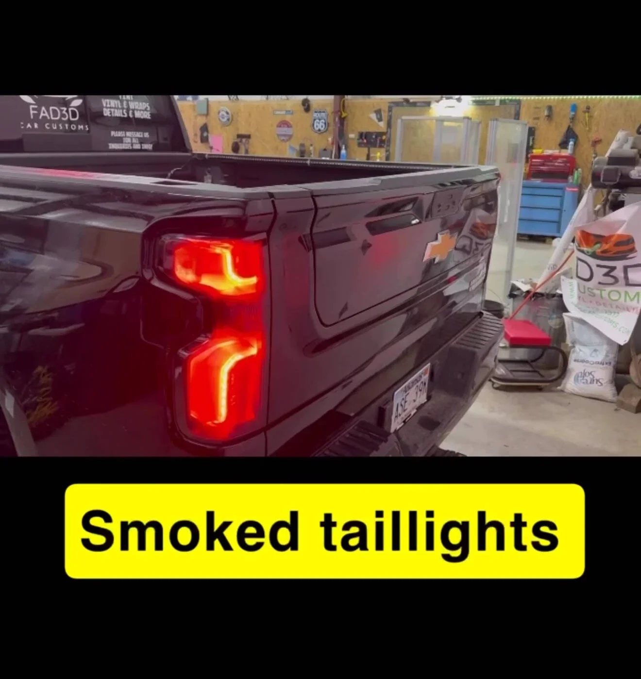 The tailgate of a black Chevrolet pickup truck with smoked taillights in a garage or workshop setting, with shelves, tools, and equipment visible in the background.