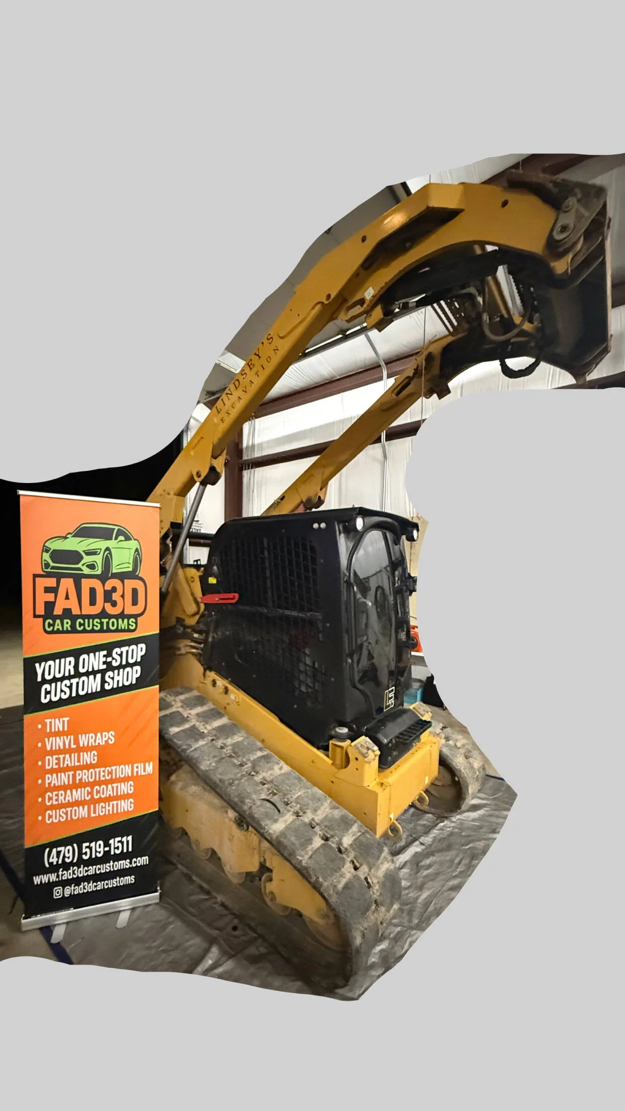 Miniature construction excavator inside a warehouse, positioned next to a promotional sign for FAD3D Car Customs.