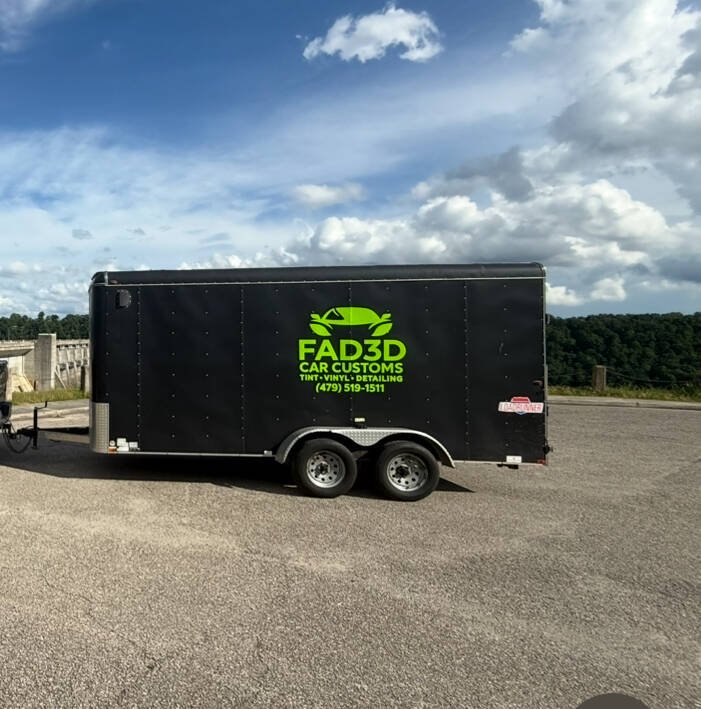 A black trailer with a green logo and text for 'FAD3D Car Customs,' including services like tint, vinyl, and detailing, parked on a gravel lot under a partly cloudy sky.