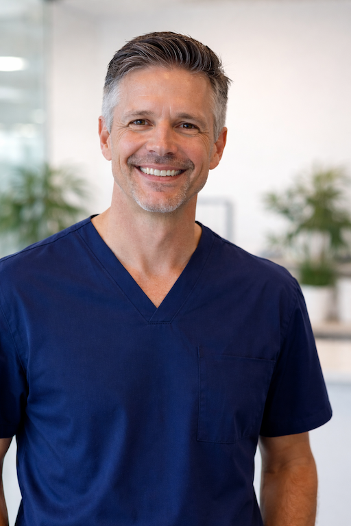 A smiling middle-aged man with salt-and-pepper hair, wearing navy medical scrubs, standing in a bright, modern medical or office space with plants in the background.