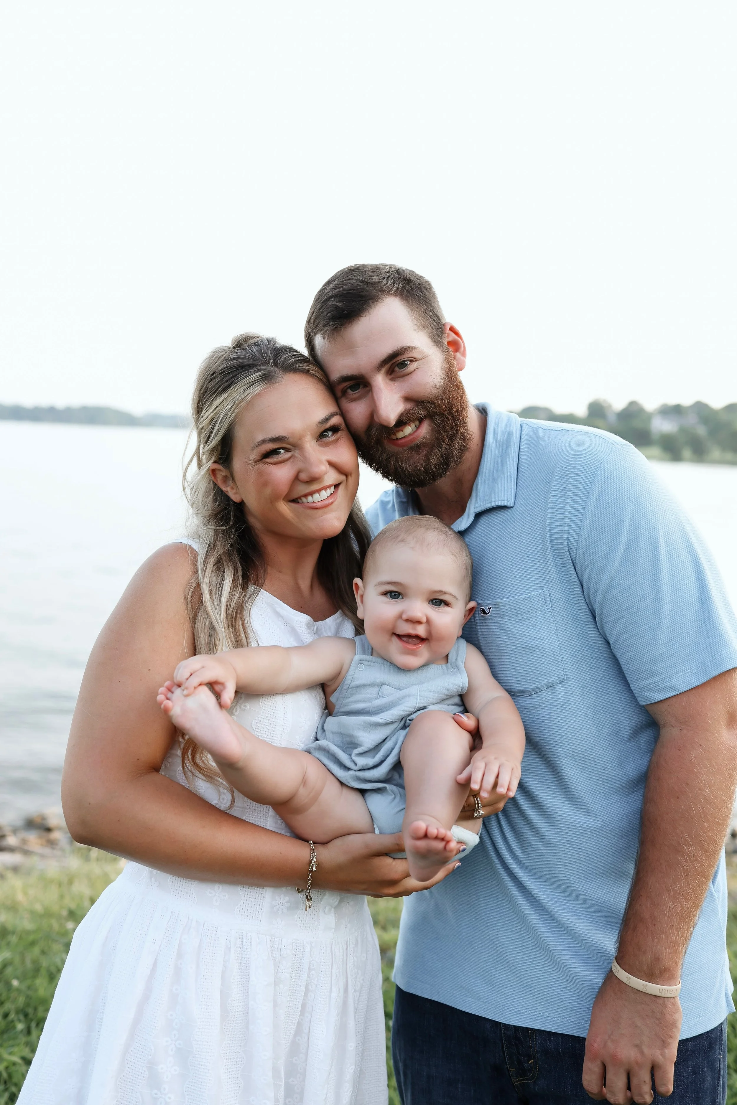 A happy family of three, a woman, a man, and a baby, standing outdoors near a body of water during daytime, smiling at the camera.