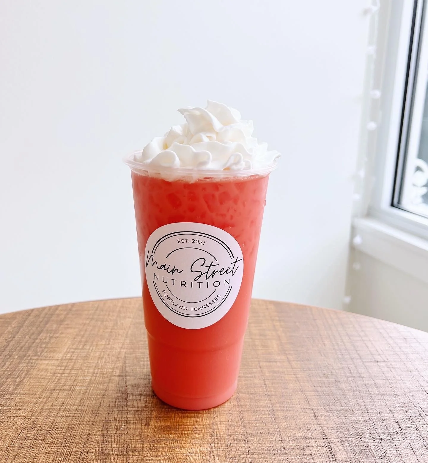 A pink drink topped with whipped cream, in a plastic cup with a white circular label that reads 'Main Street Nutrition, Portland, Tennessee, Est. 2021'.