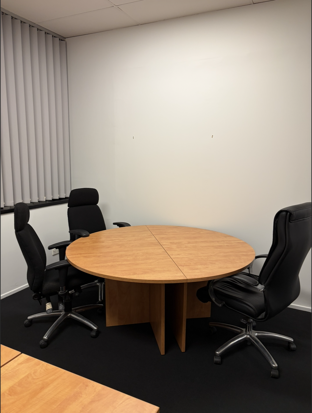 Empty office conference room with a round wooden table and three black office chairs, white walls, and vertical blinds on the window.