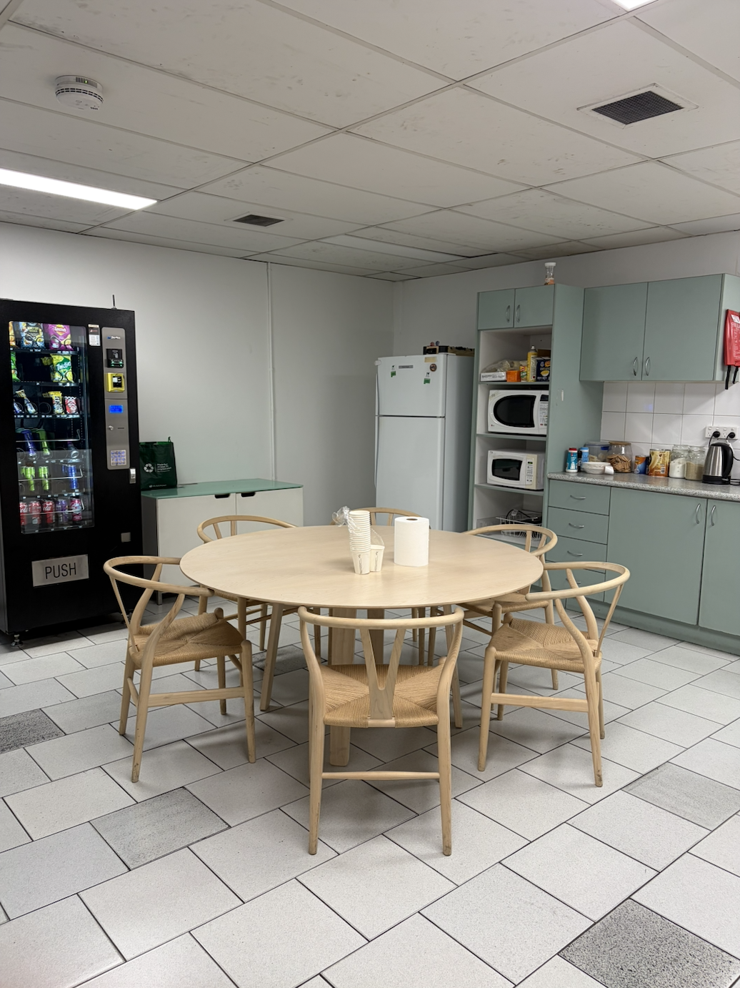 An indoor break room with a round wooden table surrounded by six matching chairs, a vending machine to the left, a white refrigerator in the background, and a kitchen area with cabinets, microwave, toaster, kettle, and various food items on the count