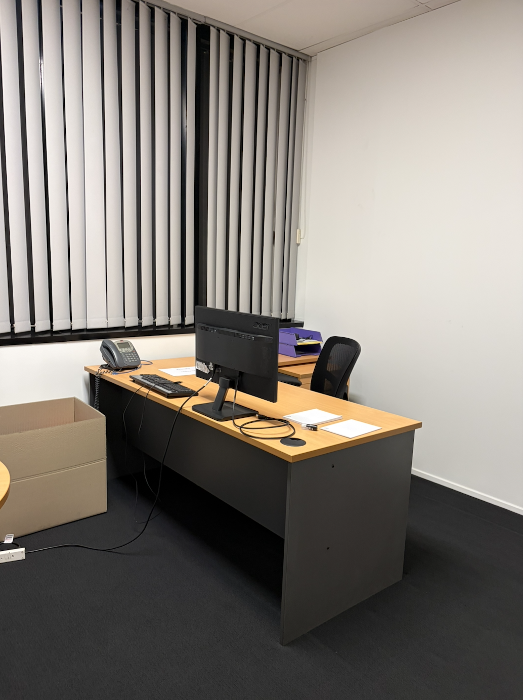 Office desk with a black computer monitor, telephone, keyboard, mouse, papers, and purple files, with a black office chair, next to a window with vertical blinds, and a cardboard box on the floor.