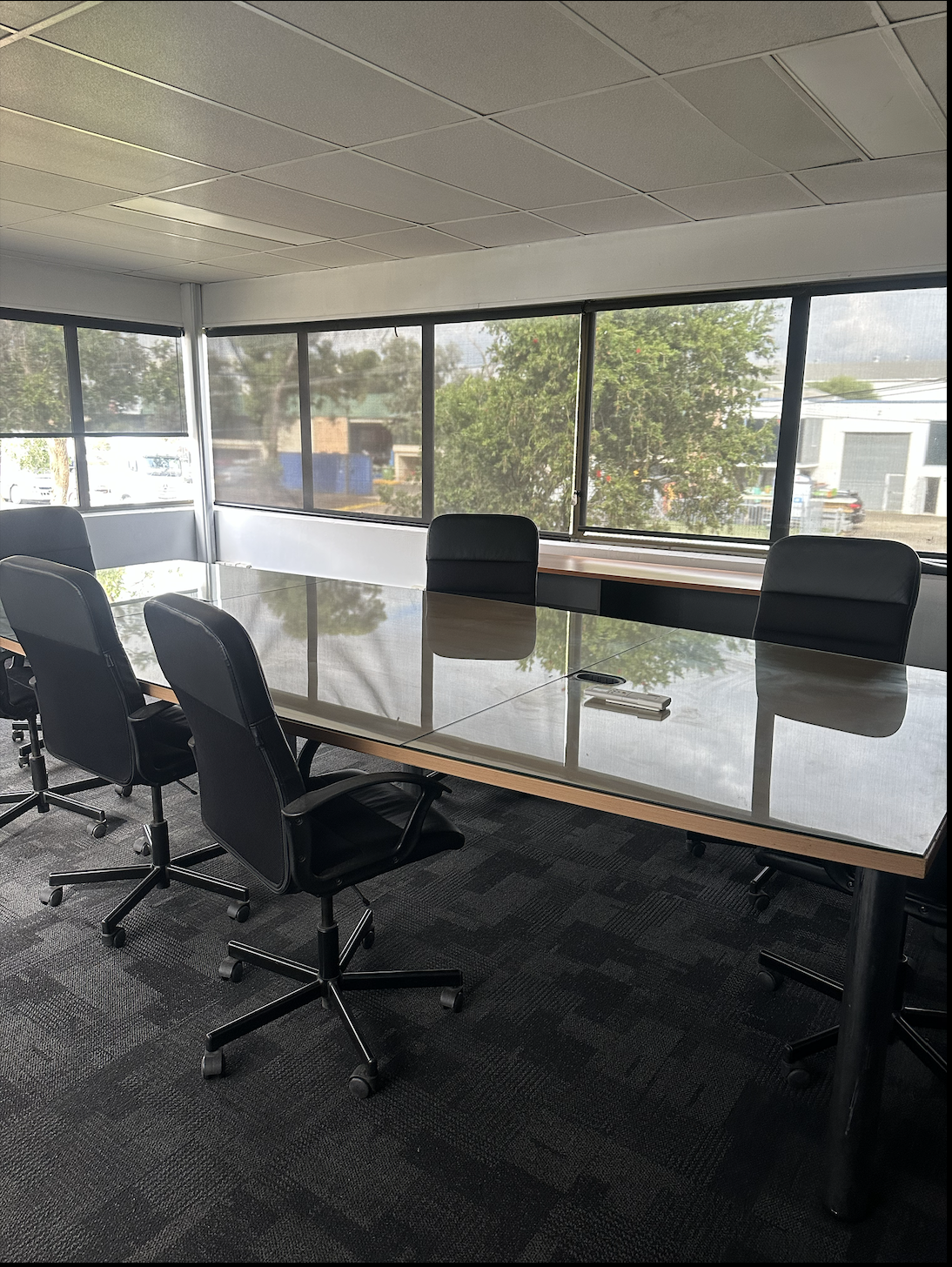An empty conference room with a long rectangular table, six black office chairs, and large windows with view of trees and buildings outside.