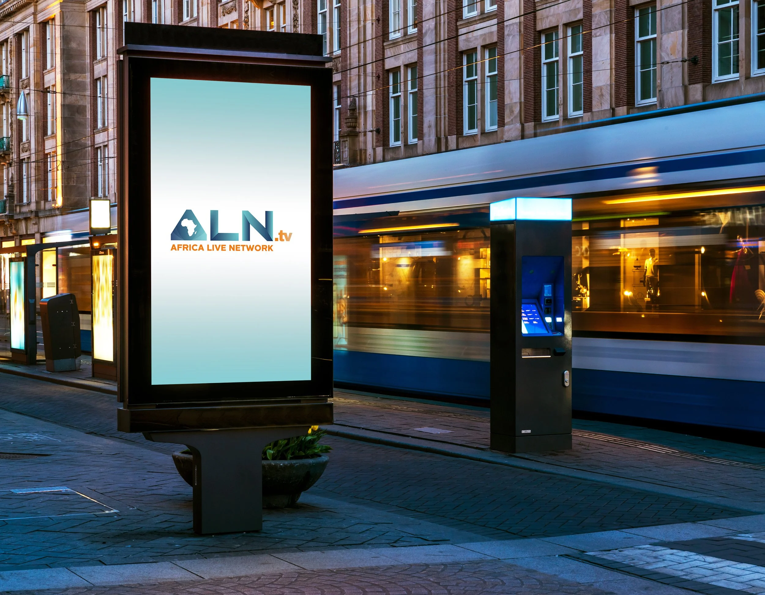 Bus stop with a digital advertisement sign displaying 'Africa Live Network' and a stylized map of Africa. The background shows a city street with a moving bus, a multi-story brick building with lit windows, and string lights overhead.