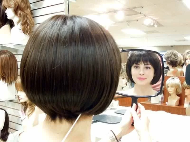 Person with short brown bob haircut holding a mirror, reflected image shows a woman with a short bob hairstyle inside a wig shop, with mannequin heads wearing wigs on the wall in the background.