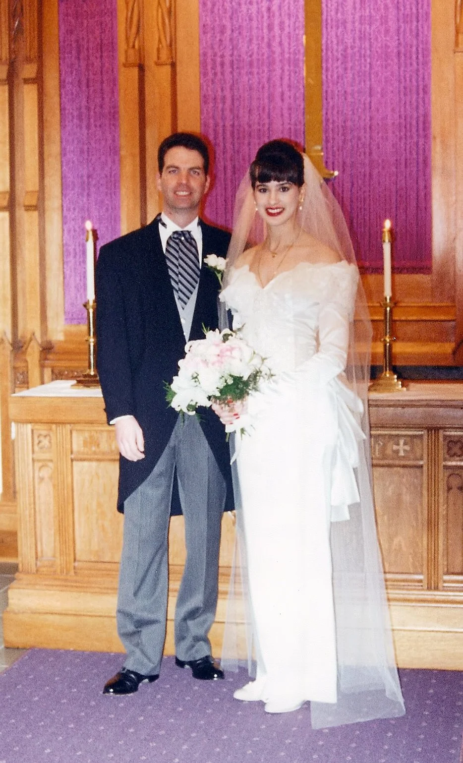 A wedding couple standing together inside a church with purple and wooden altar backdrop. The bride is wearing a white wedding dress with long sleeves, a veil, and holding a bouquet of pink and white flowers. The groom is dressed in a black tuxedo with a white shirt, striped tie, and gray trousers.
