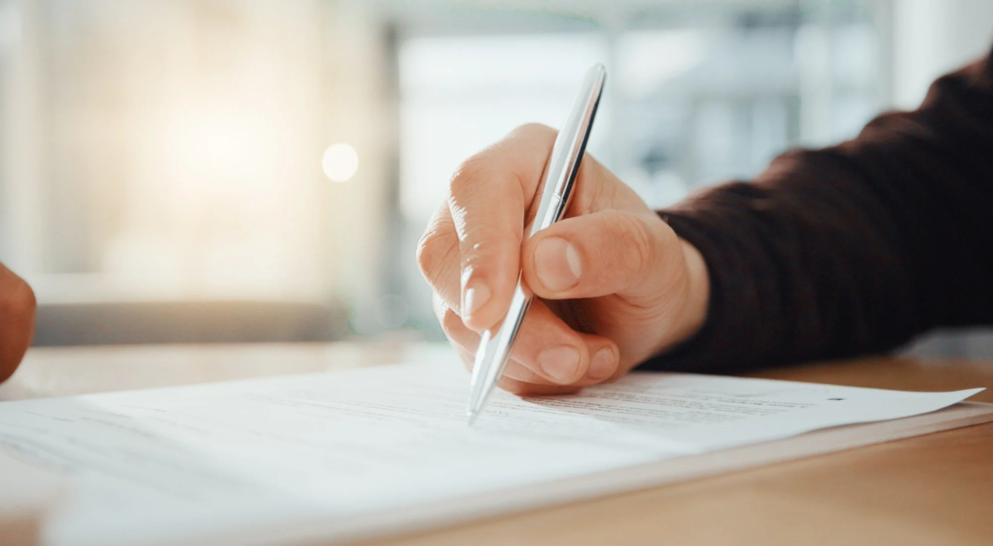 Close-up of a person holding a silver pen and writing on lined paper on a wooden table.