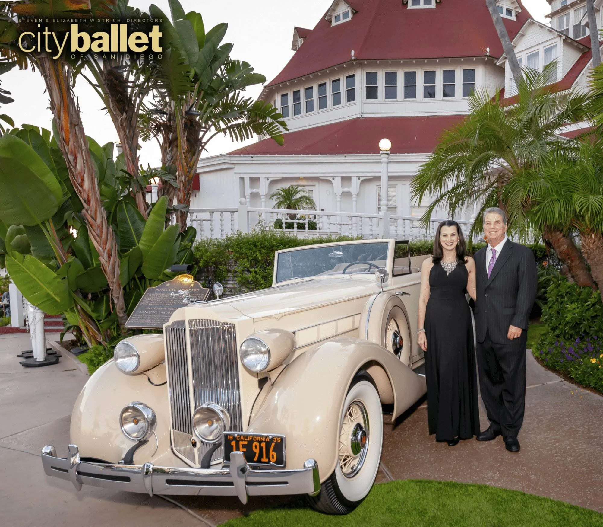 A vintage cream-colored Rolls-Royce car with a California license plate, parked in front of a white house with a red roof, surrounded by lush greenery and tall palm trees. Two people in formal attire, a woman and a man, stand next to the car.