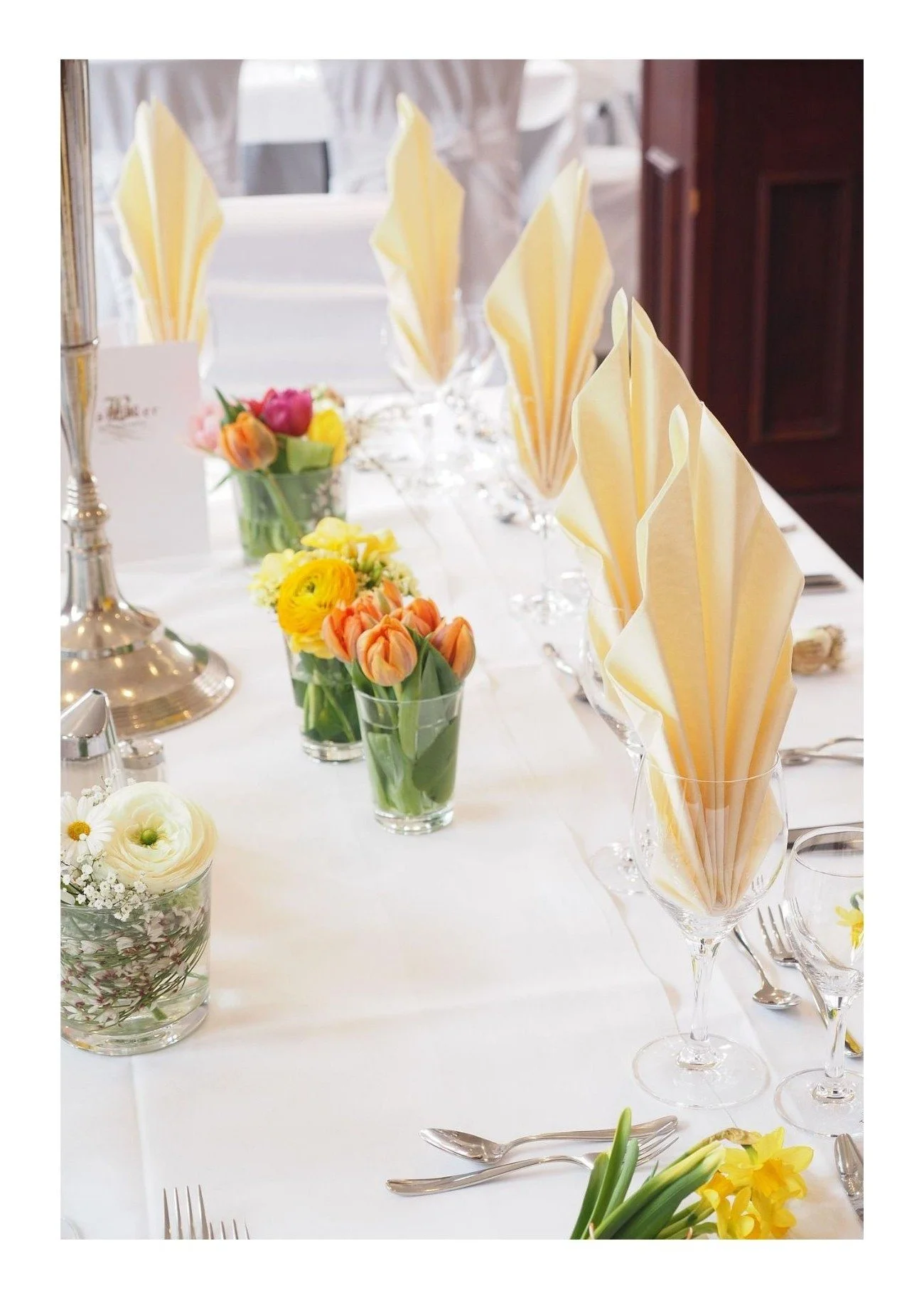 Elegant table setting with yellow and white napkins folded in a fan shape in wine glasses, adorned with floral centerpieces including tulips, roses, and other colorful flowers, and a white tablecloth.