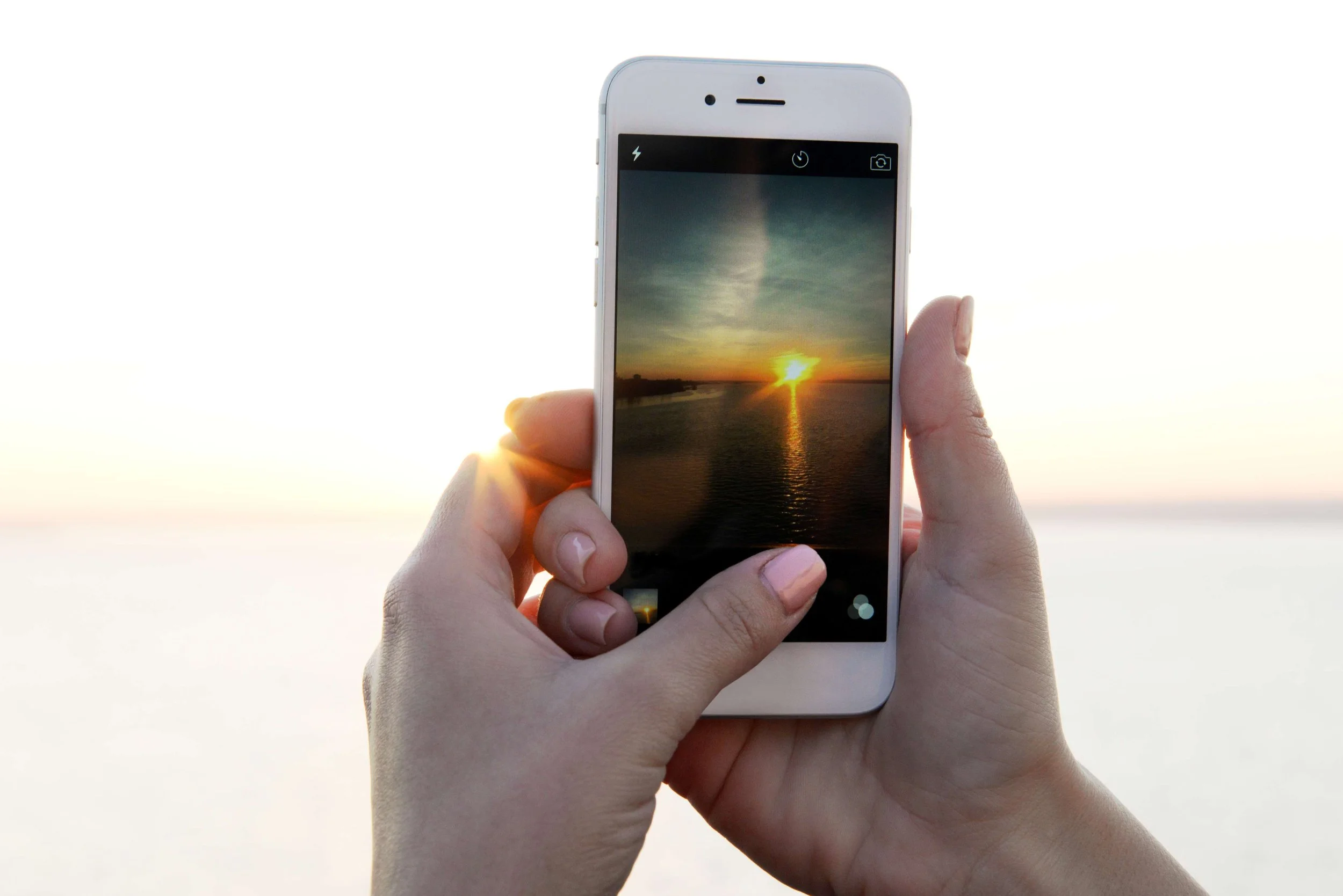 Person holding a smartphone capturing a photo of a sunset over a body of water.