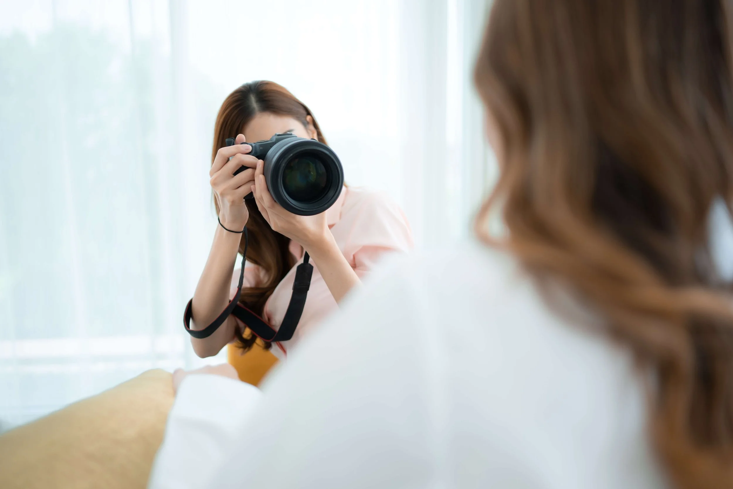 A woman taking a photograph of another person with a digital camera indoors near a window.