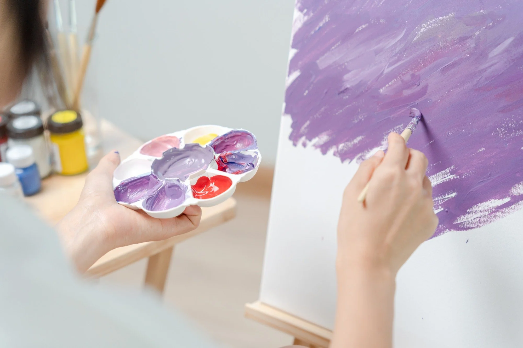 Person mixing purple and red paint on a palette while painting a purple background on a canvas.