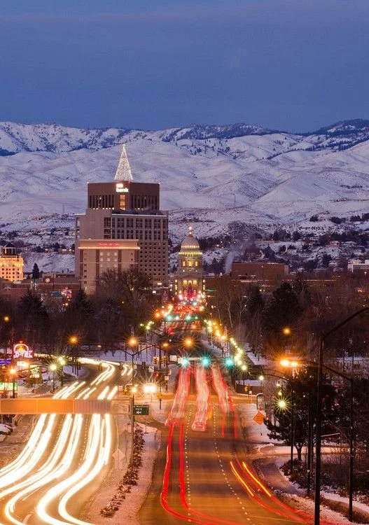 A city street at night with light trails from moving vehicles, with tall buildings and snow-covered mountains in the background.