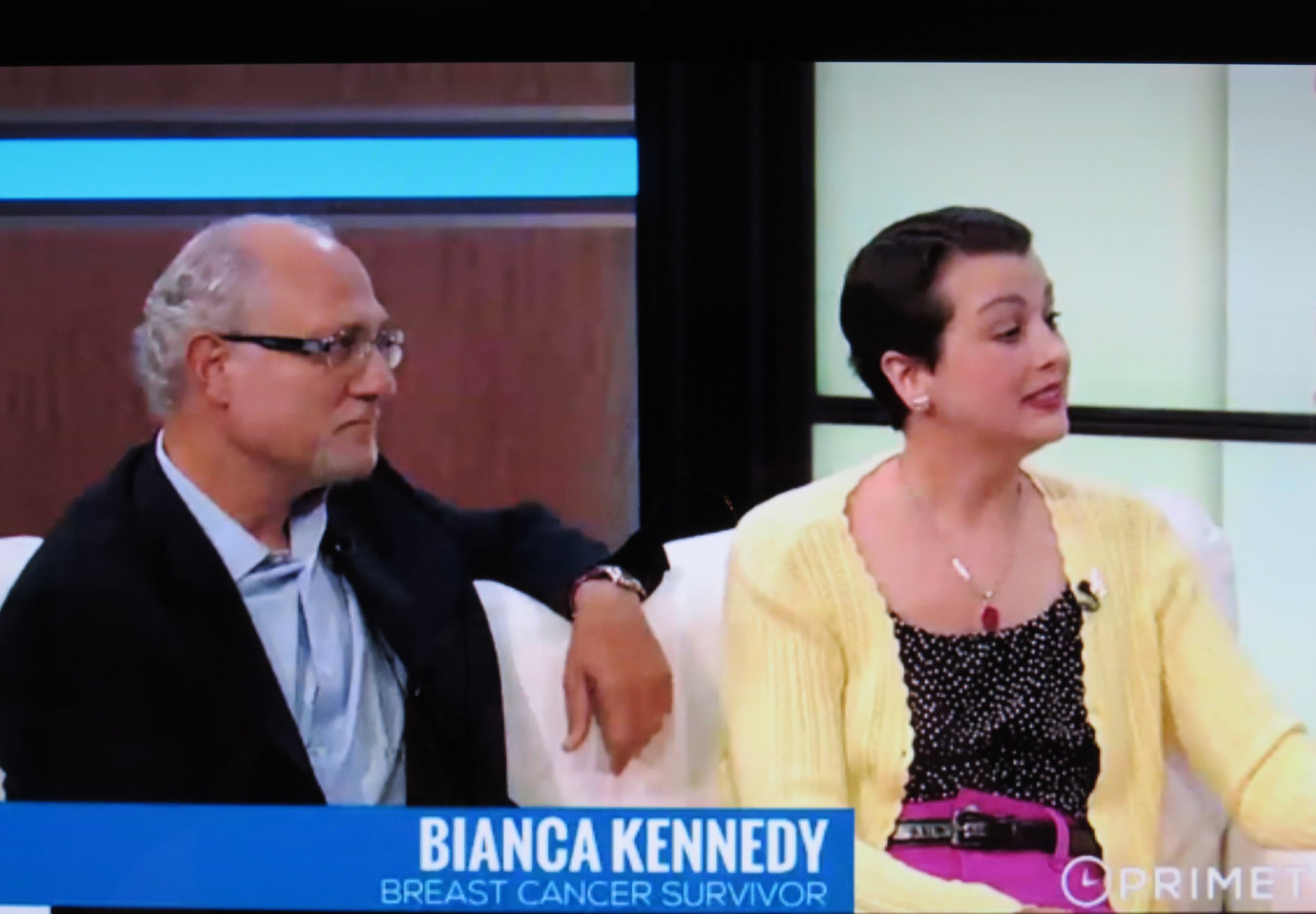 A television screen showing a news segment featuring a woman in a yellow cardigan and a man with glasses, with a caption reading "Bianca Kennedy, Breast Cancer Survivor."