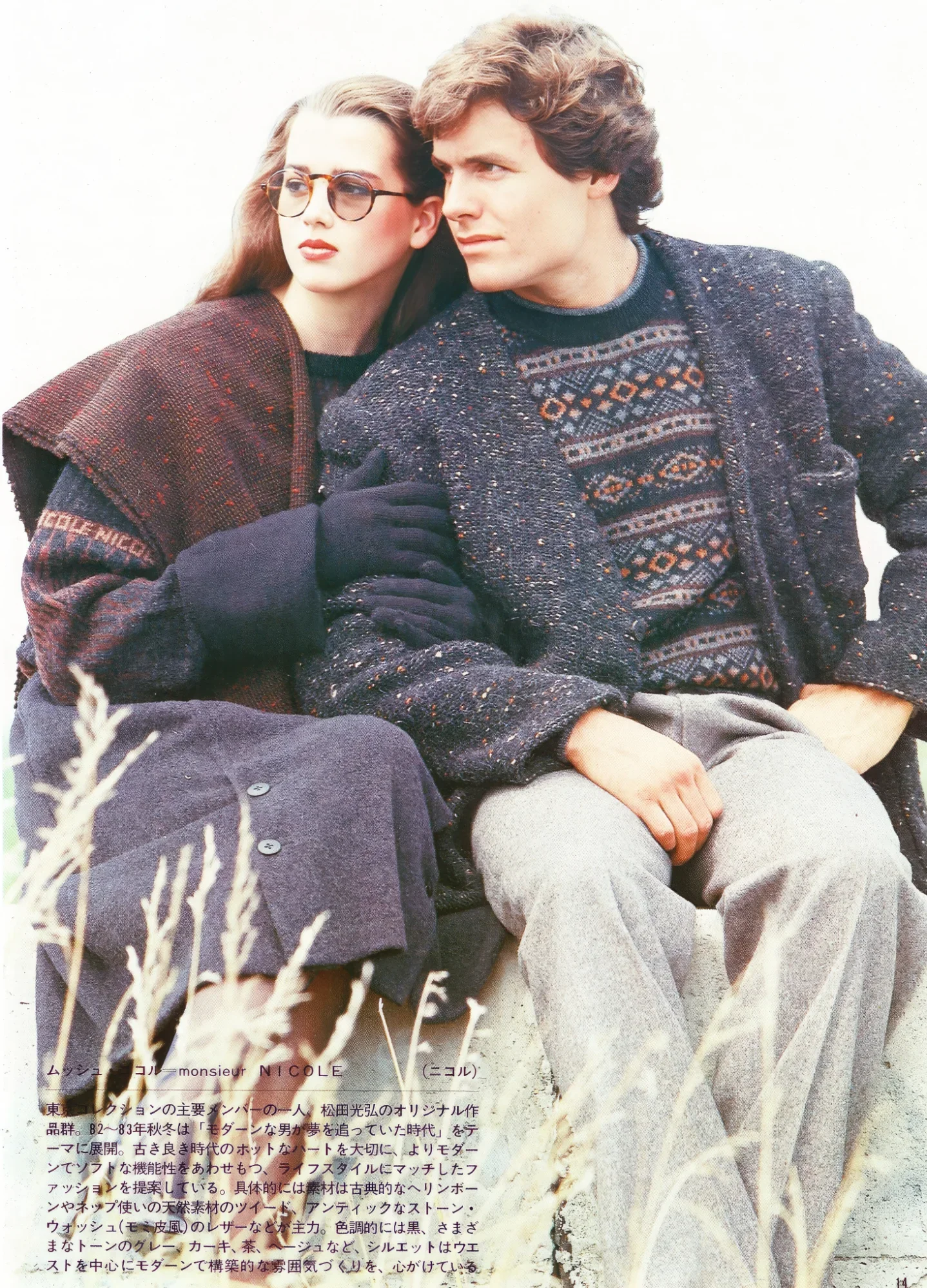 Two young adults, a woman with glasses and long hair, and a man with curly hair, sit closely together outdoors against a white background. They are dressed in warm, vintage-style sweaters and jackets, with muted color tones and textured fabrics.