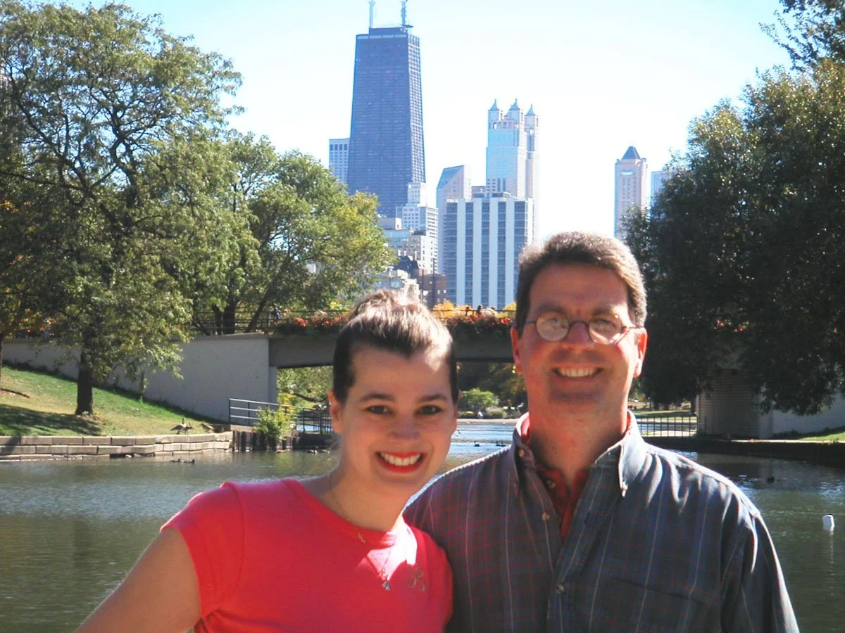 Two people smiling in front of a Chicago cityscape with tall buildings and a river, with trees on the banks of the river.