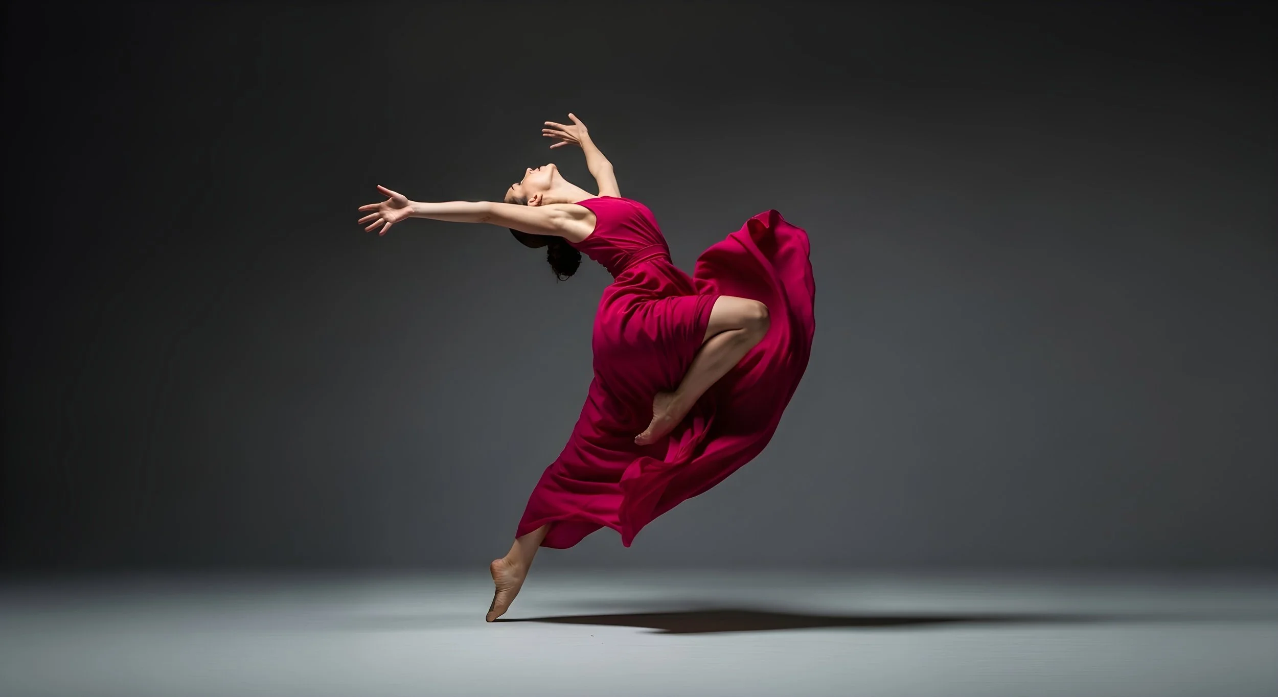 A dancer performing an striking pose in a studio with a dark gray background, wearing a flowing red dress.