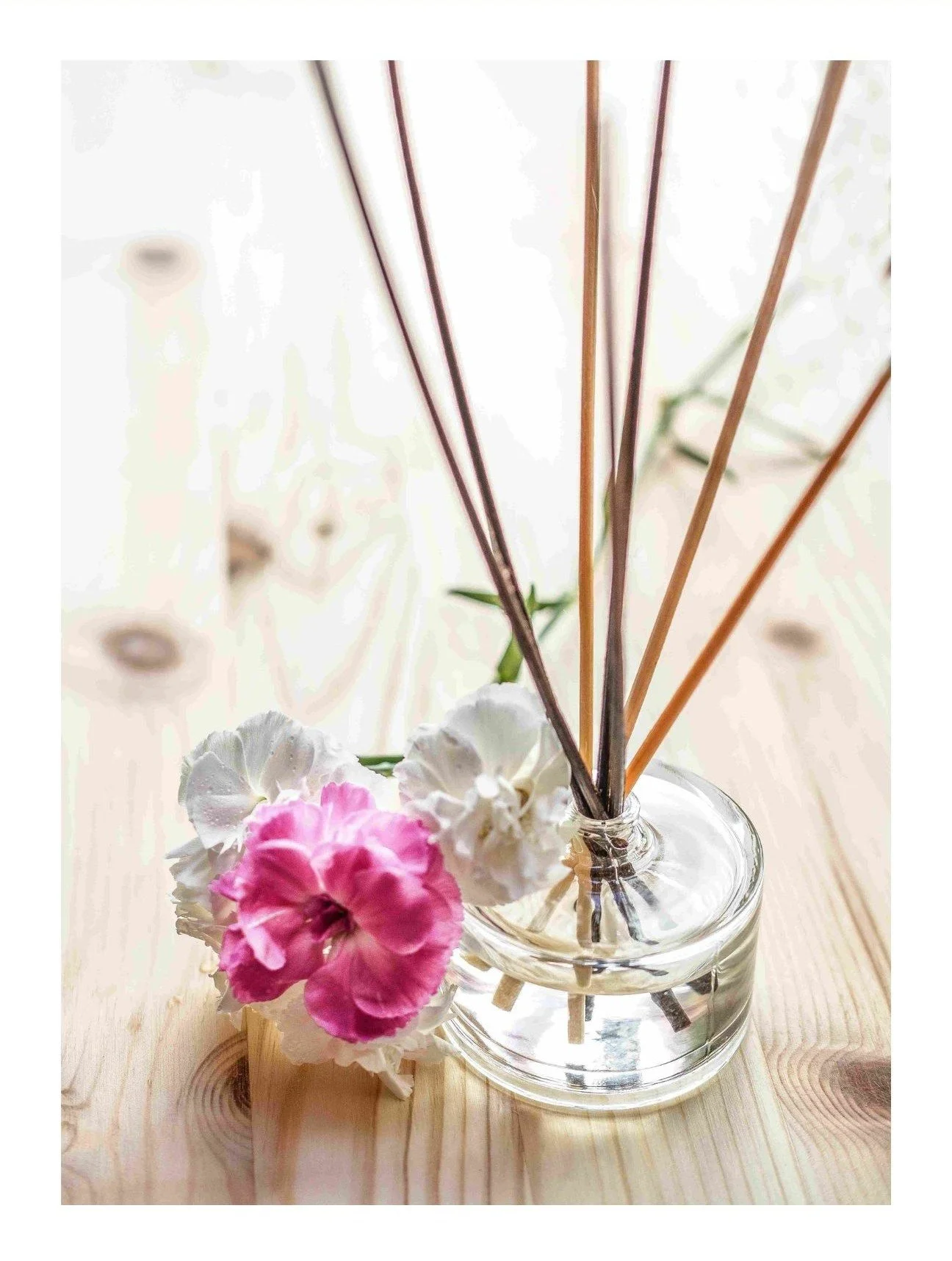 A glass vase with pink and white flowers, and diffusers with long sticks, on a wooden surface.