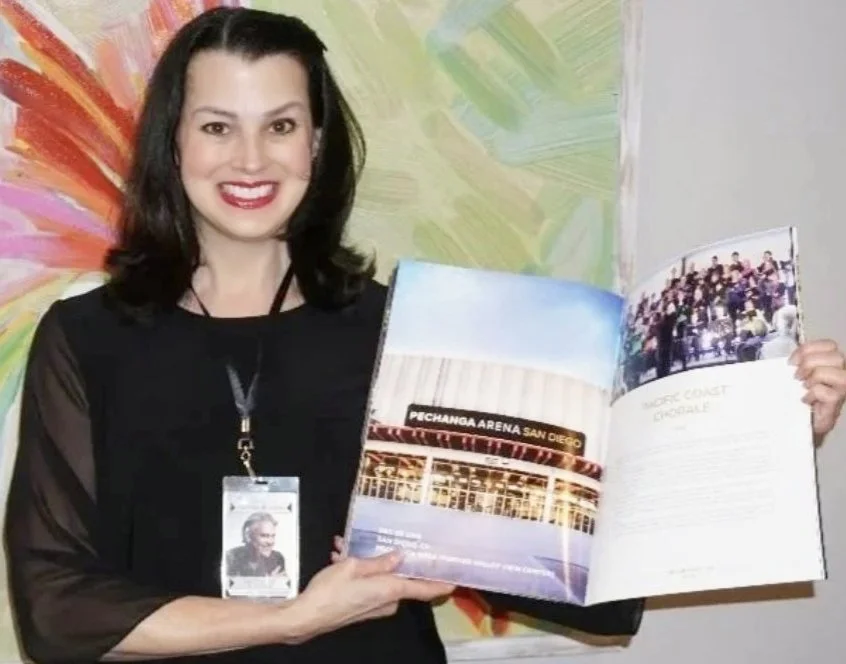 A woman with dark hair, smiling, wearing a black top with sheer sleeves, holding an open concert tour program showing an image of a stadium and a choir photo.