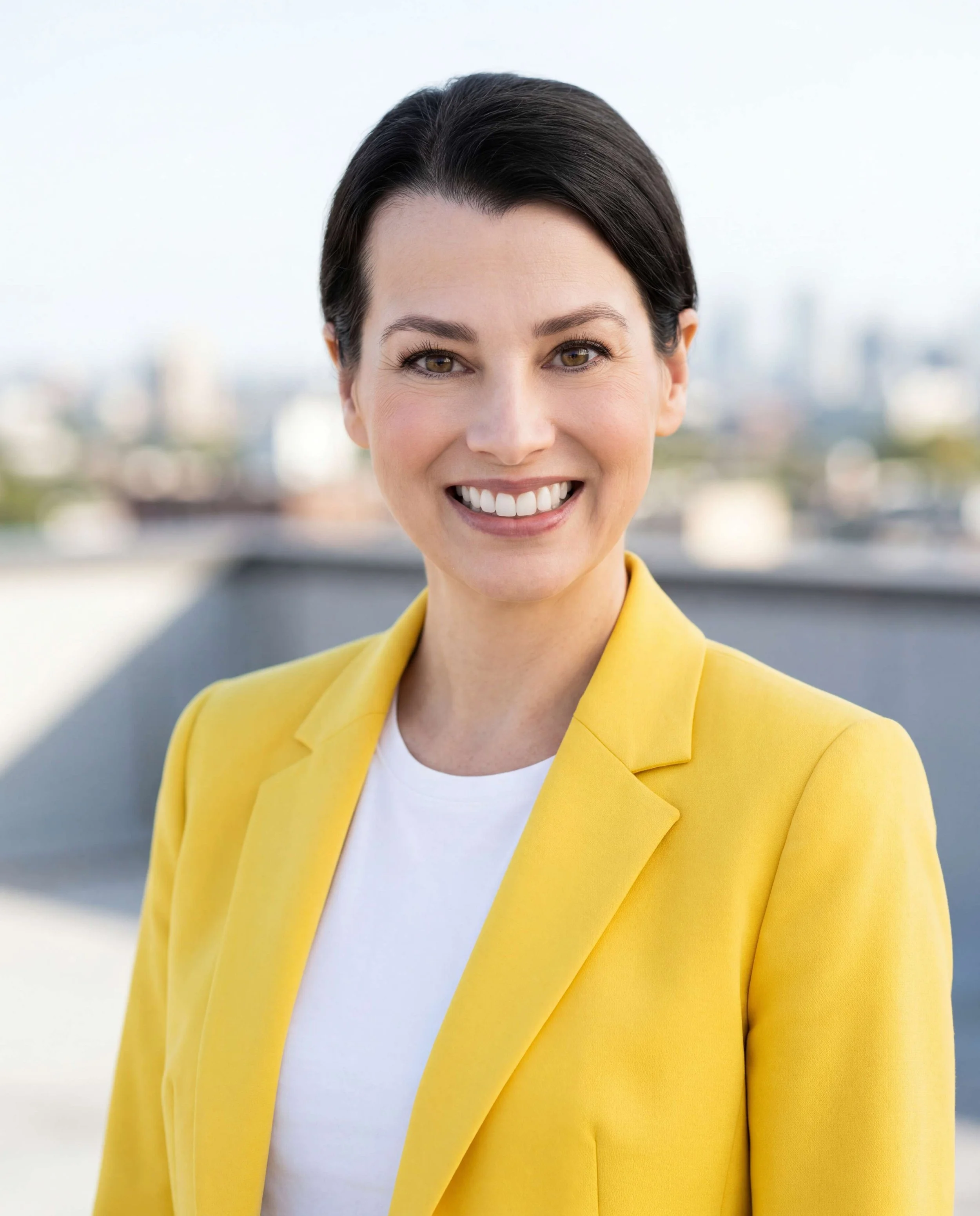 A woman with dark hair smiling, wearing a yellow blazer over a white top, with a blurred cityscape background.