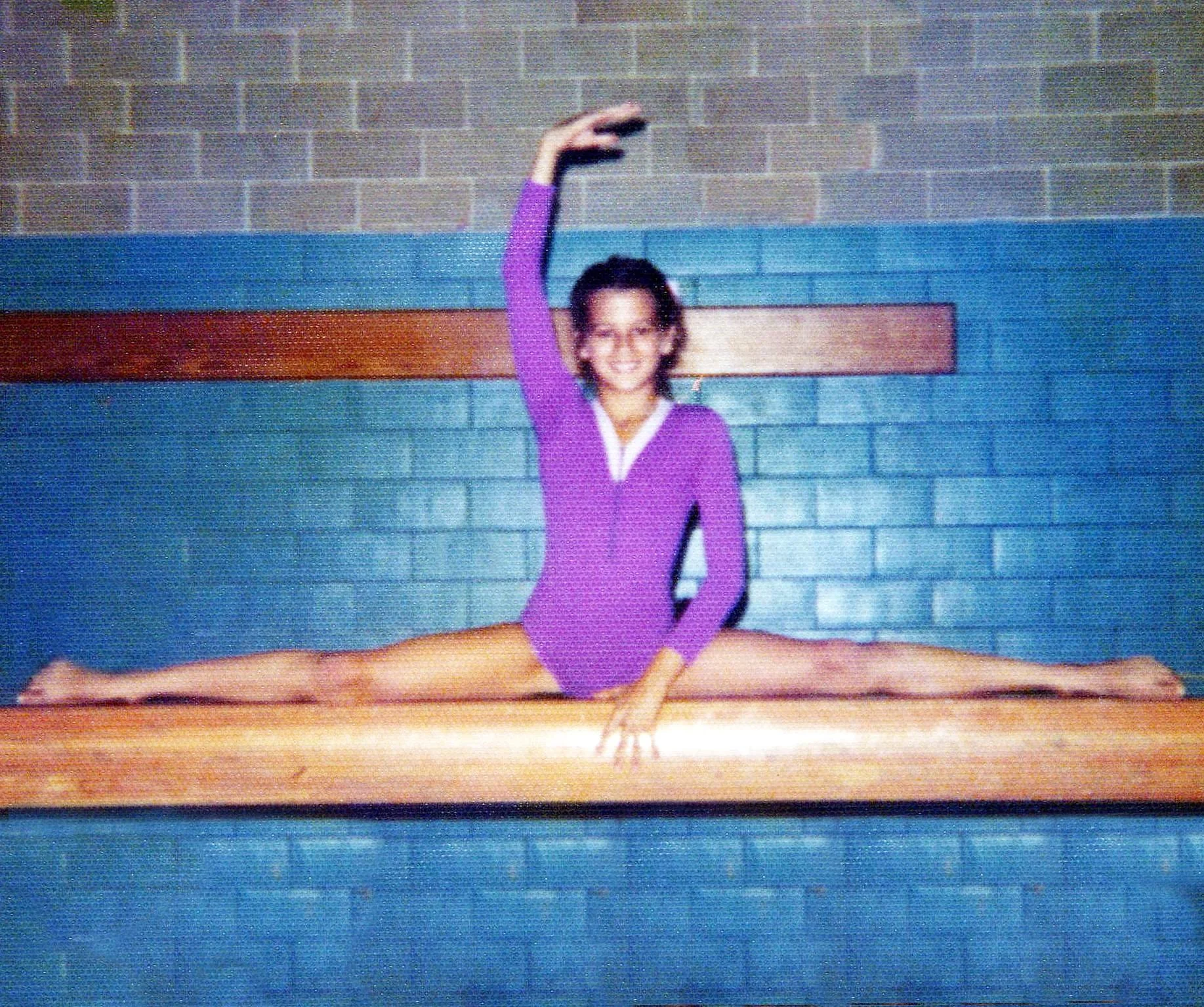 A young girl in a purple leotard sitting on a balance beam with her right arm raised, smiling, in front of a blue brick wall with a horizontal wooden beam.