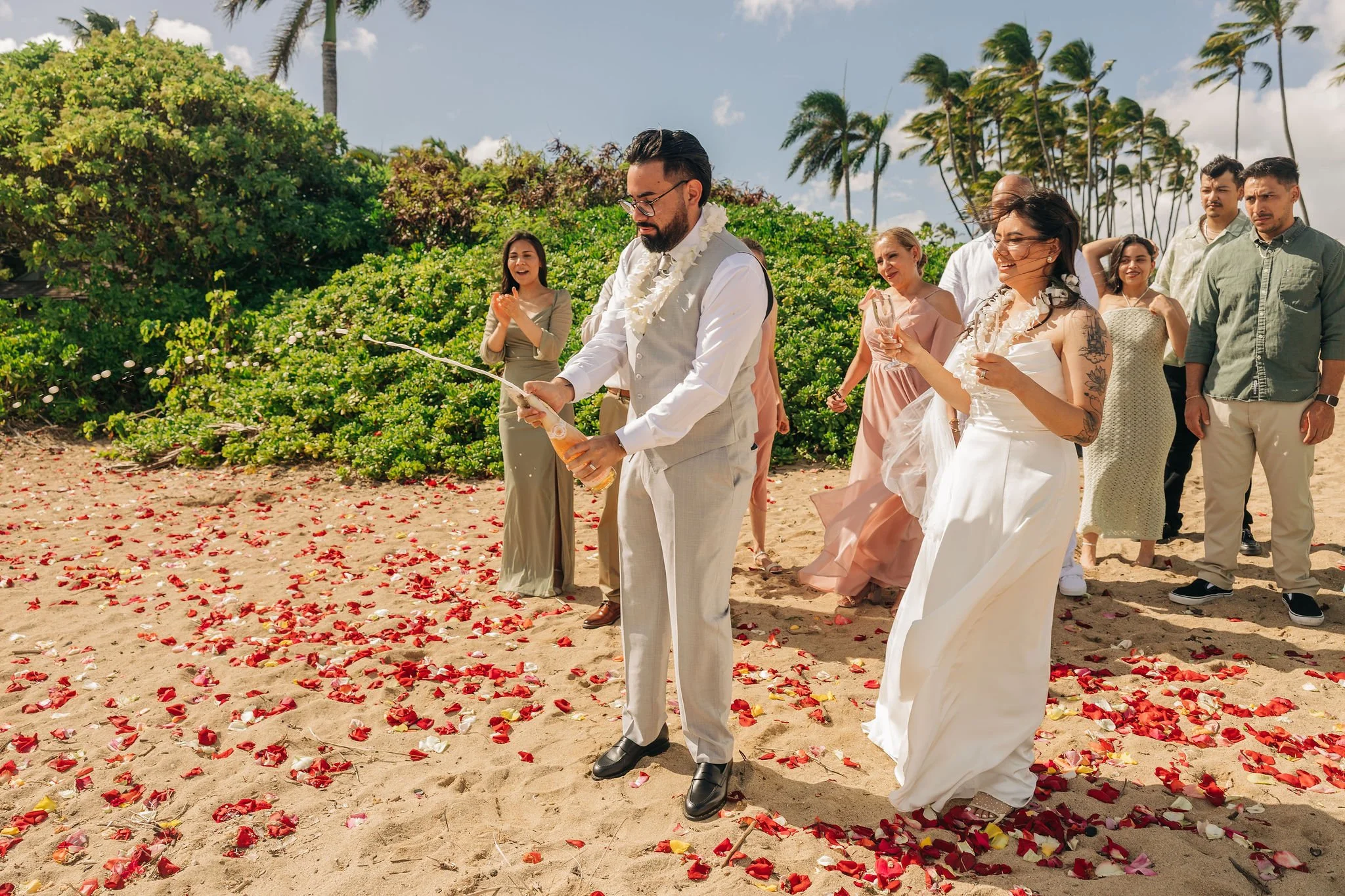 A group of people celebrating a wedding on a beach, with a groom opening a bottle of champagne, surrounded by flower petals and palm trees.