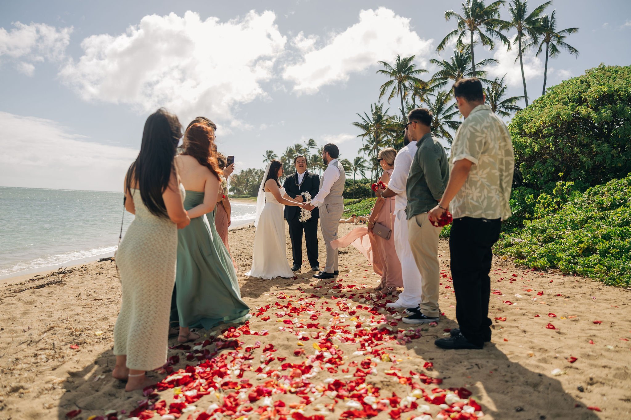 A beach wedding ceremony with a group of people standing on a sandy beach, exchanging vows under a partly cloudy sky, with palm trees and the ocean in the background, and red flower petals scattered on the ground.