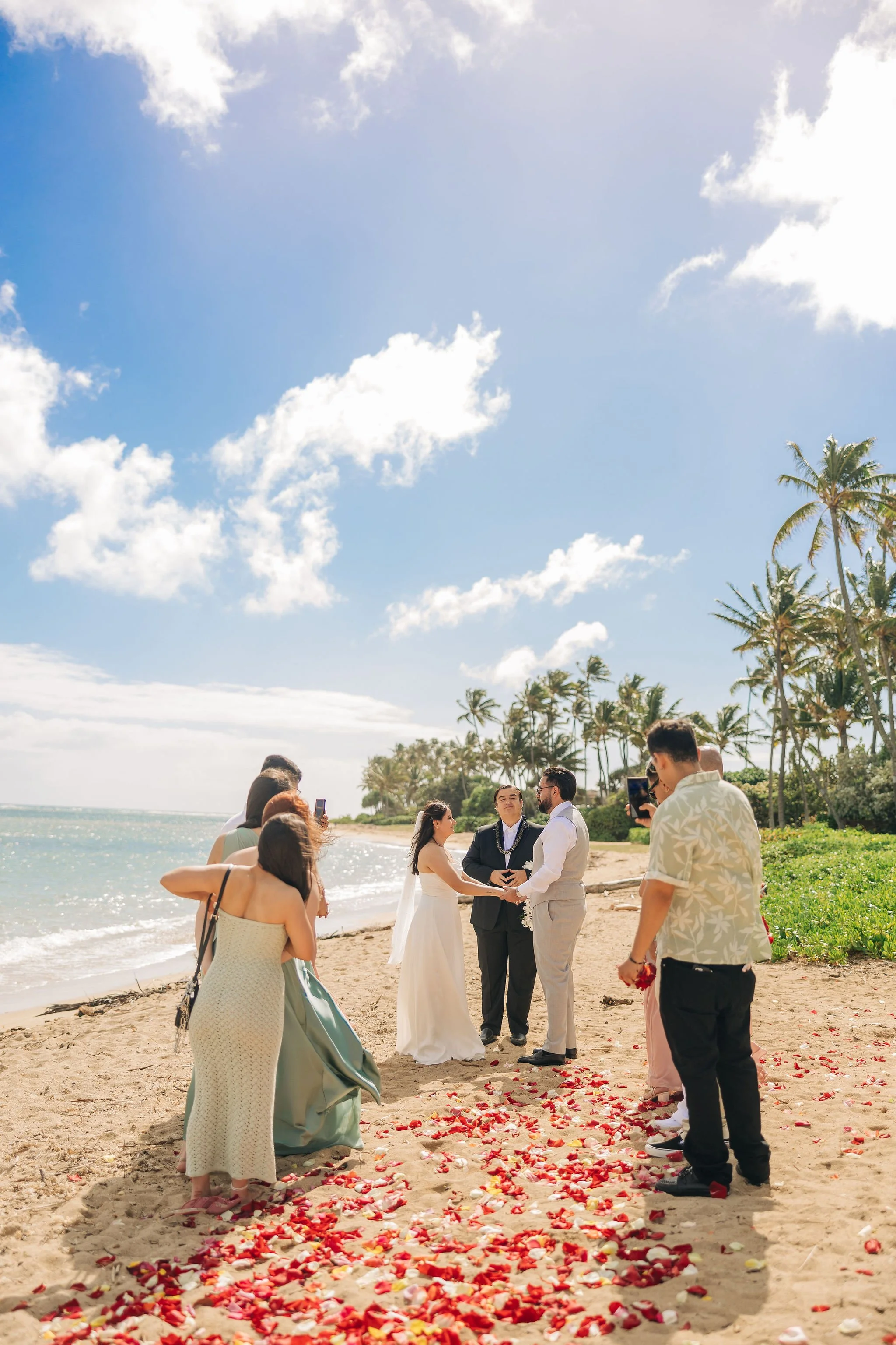 A beach wedding ceremony taking place under a bright blue sky with scattered clouds, palm trees in the background, and flower petals scattered on the sand, with a bride and groom holding hands and exchanging vows surrounded by friends and family.