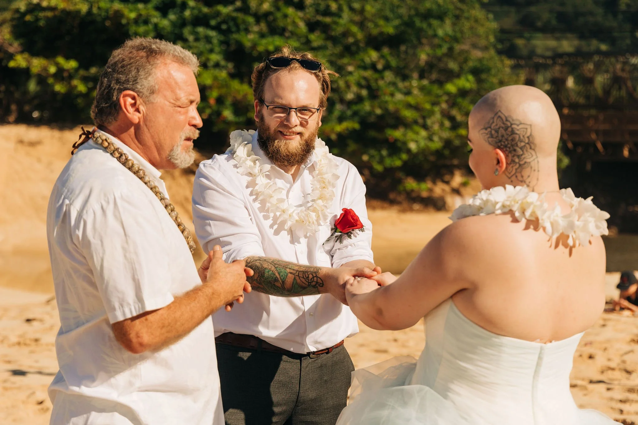 A wedding ceremony on a beach with three people: an officiant, a groom, and a bride with a shaved head and tattoos, all holding hands and smiling.
