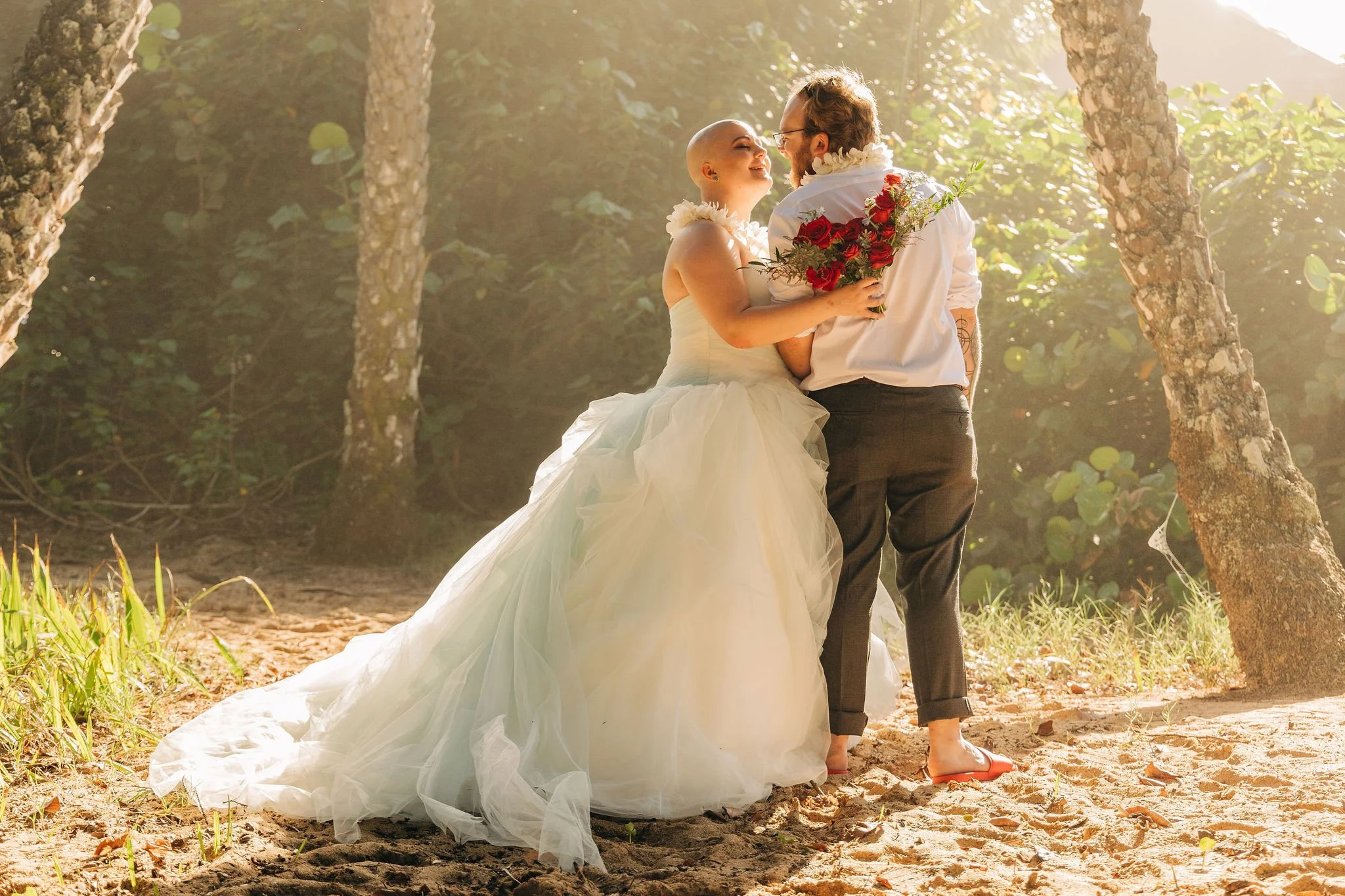 A bride and groom share a moment outdoors among trees, with the bride holding a bouquet of red roses and the groom in a white shirt and dark pants, both smiling at each other.