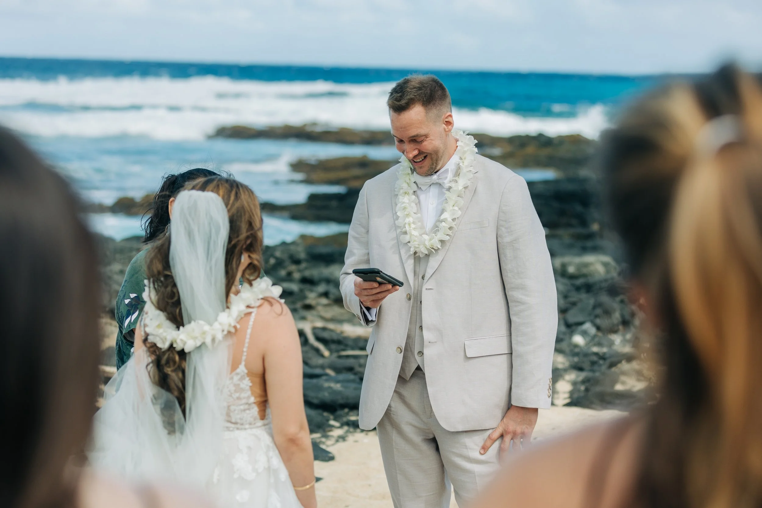 A wedding ceremony taking place on a beach with the groom reading vows, surrounded by guests, near the ocean with waves and rocks in the background.