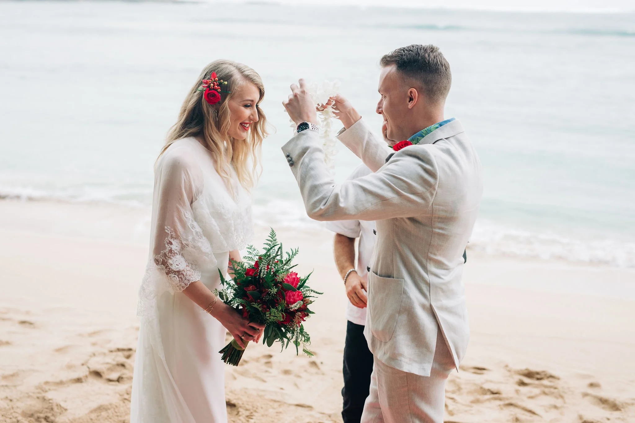 A couple getting married on the beach, with the groom placing a garland around the bride's neck, while the bride holds a bouquet of red and green flowers, and both wear light-colored clothing with floral accents.